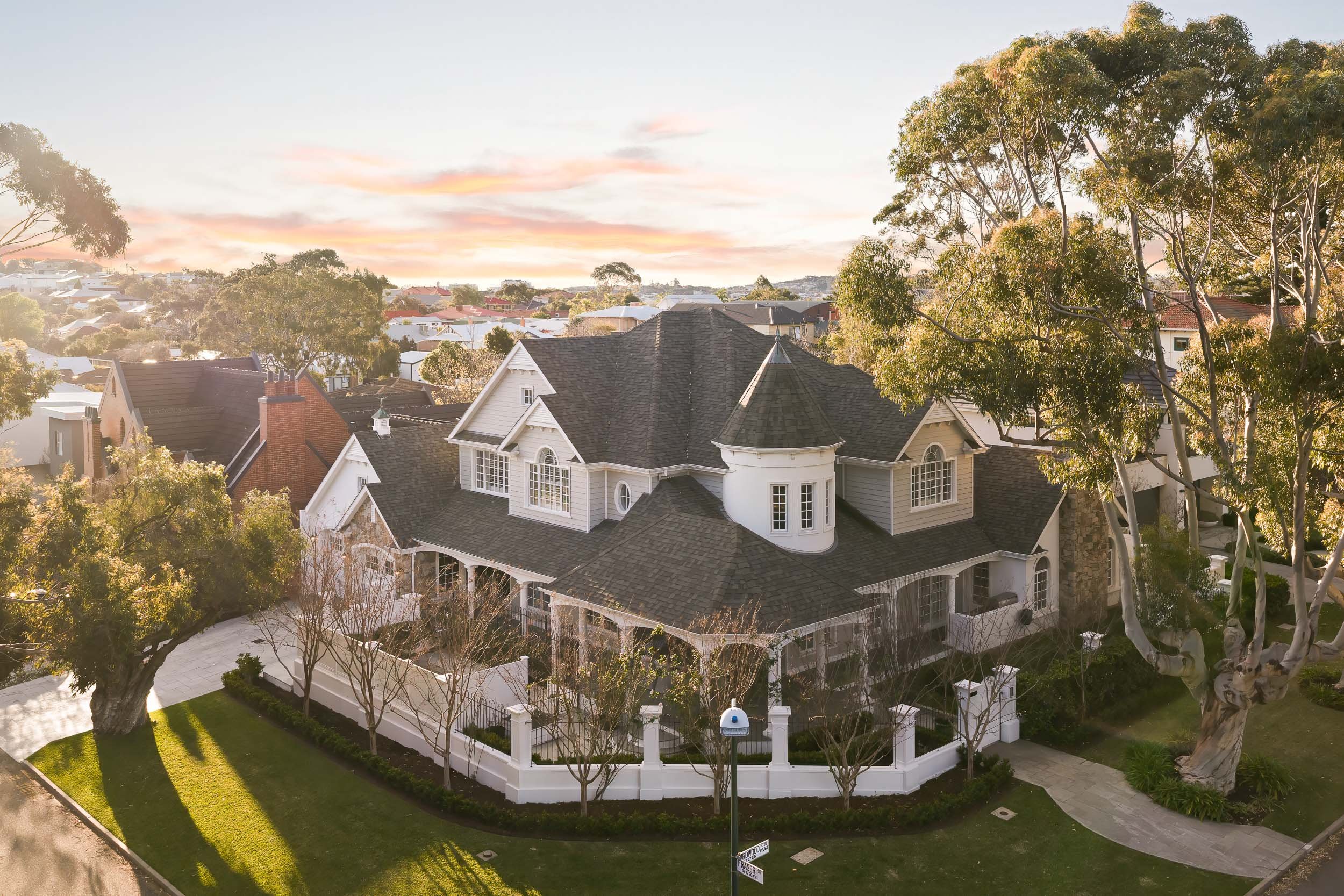 Aerial view of a large traditional home with gabled roofs, a turret and mature trees at sunset.