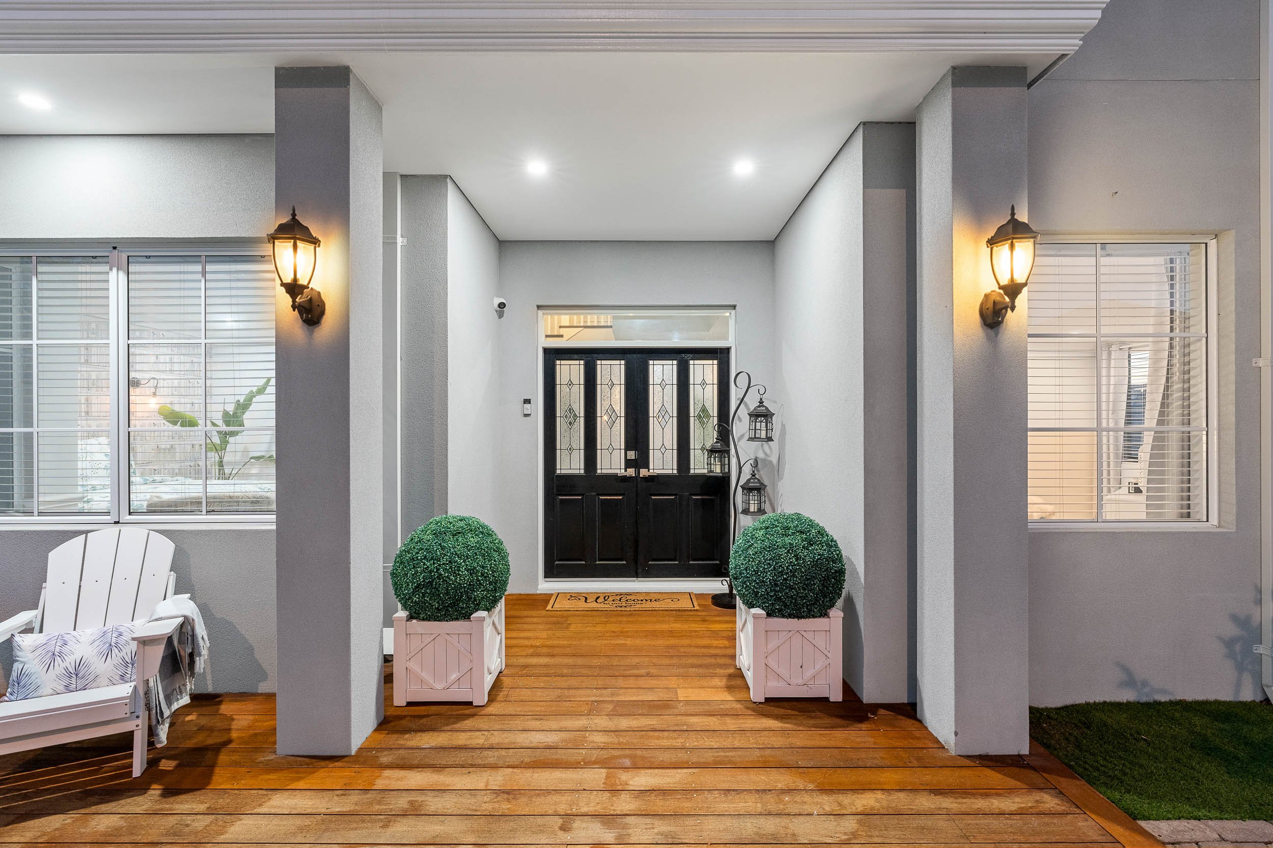 Sheltered entrance porch with wall lanterns, paired potted shrubs and a dark front door.