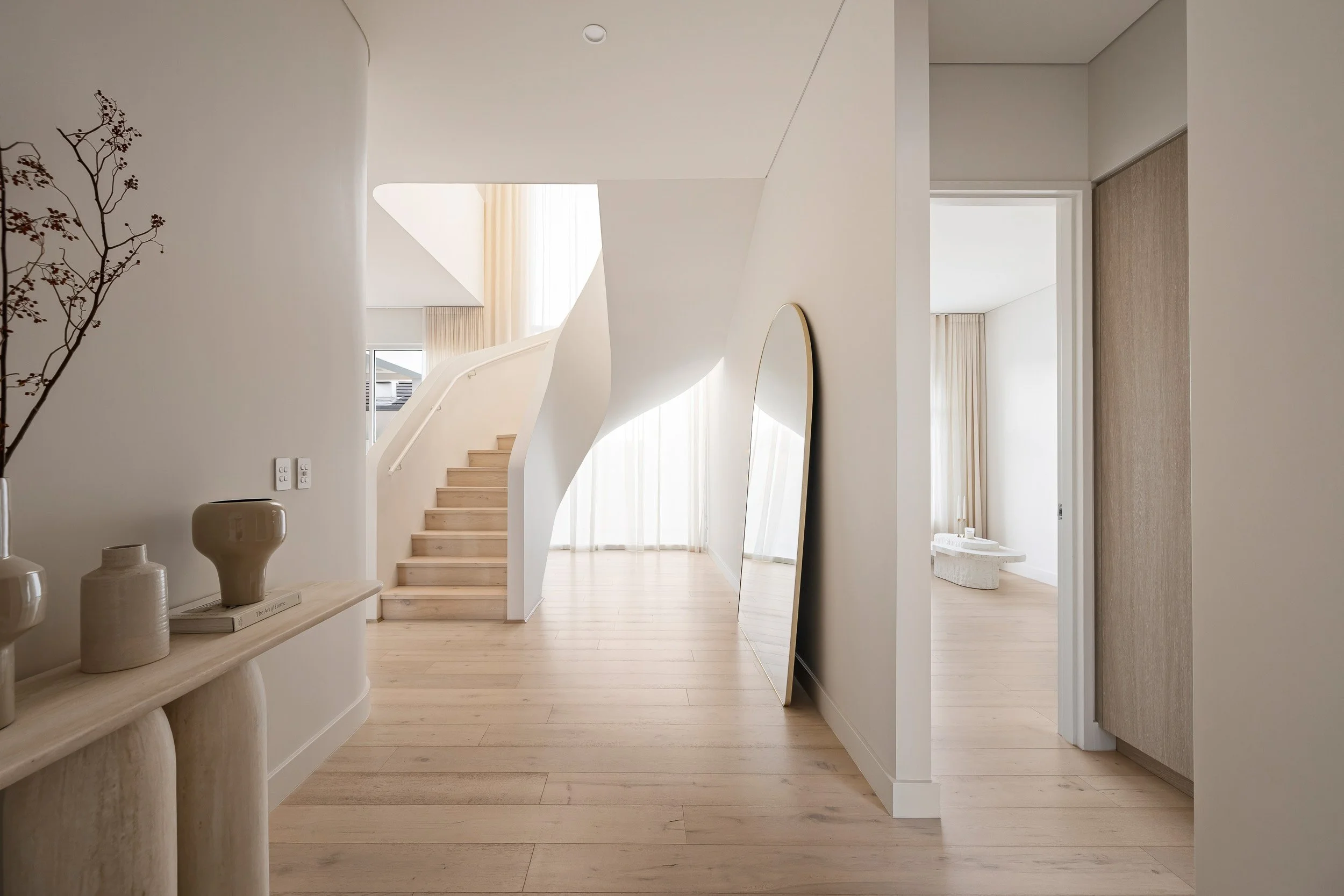 Sunlit entrance with pale timber floors, a sculptural curved staircase and an arched mirror.