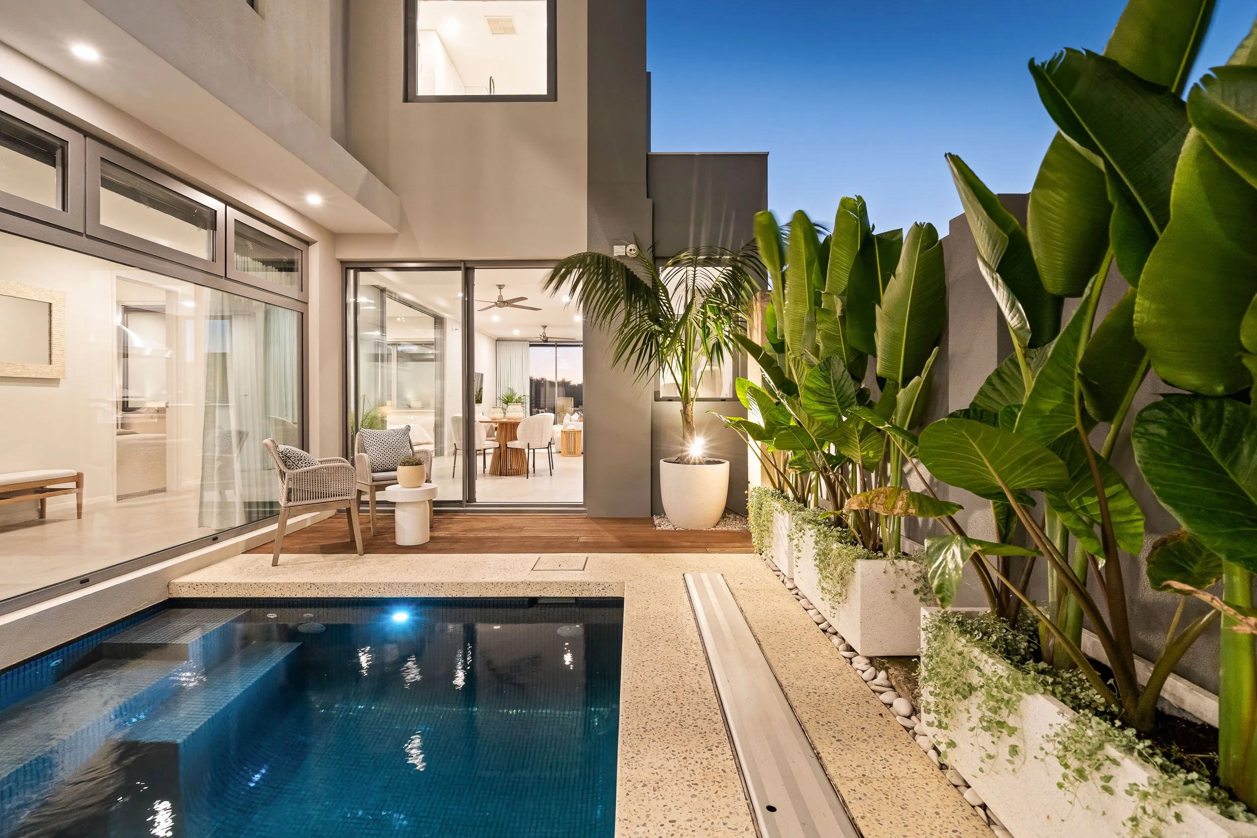 Private pool courtyard with tropical planting and a brightly lit contemporary home at dusk.