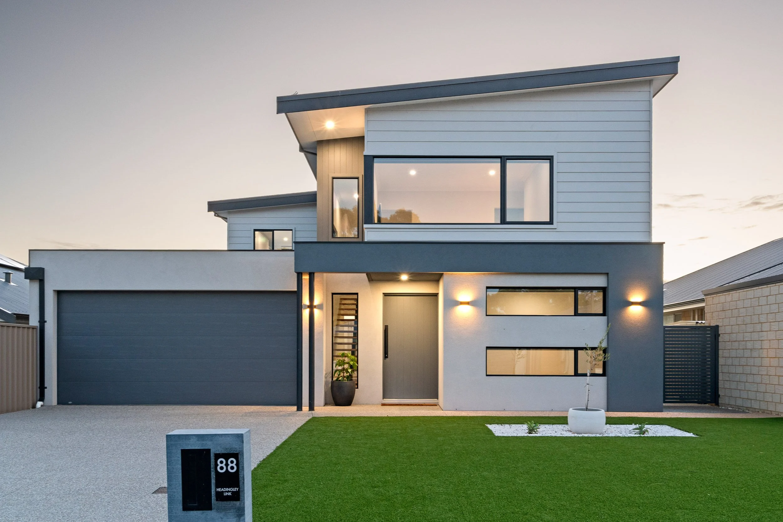 Two-storey home with charcoal cladding, warm entrance lighting and a landscaped front lawn.