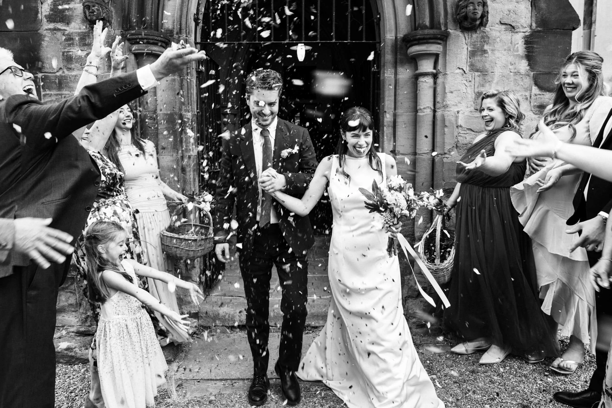 Bride and groom walk down the confetti line as their family and friends smile at them and throw confetti in the air.