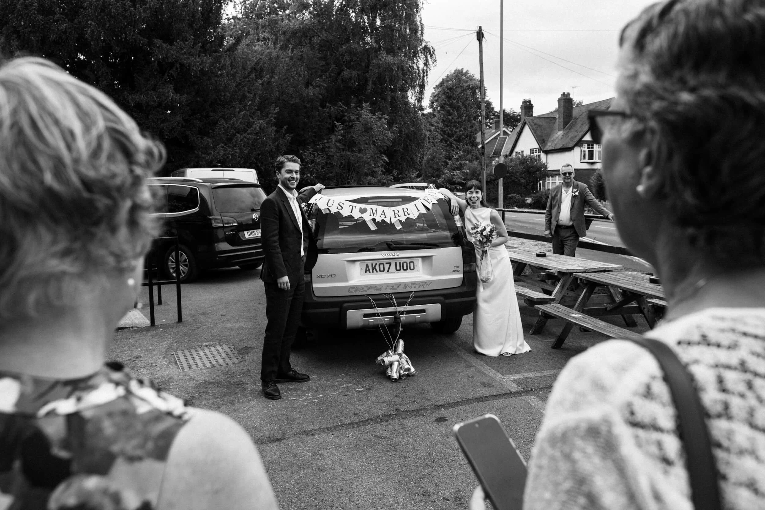 Bride and groom lean on a car that shows the sign 'just married' and has cans attached to it.