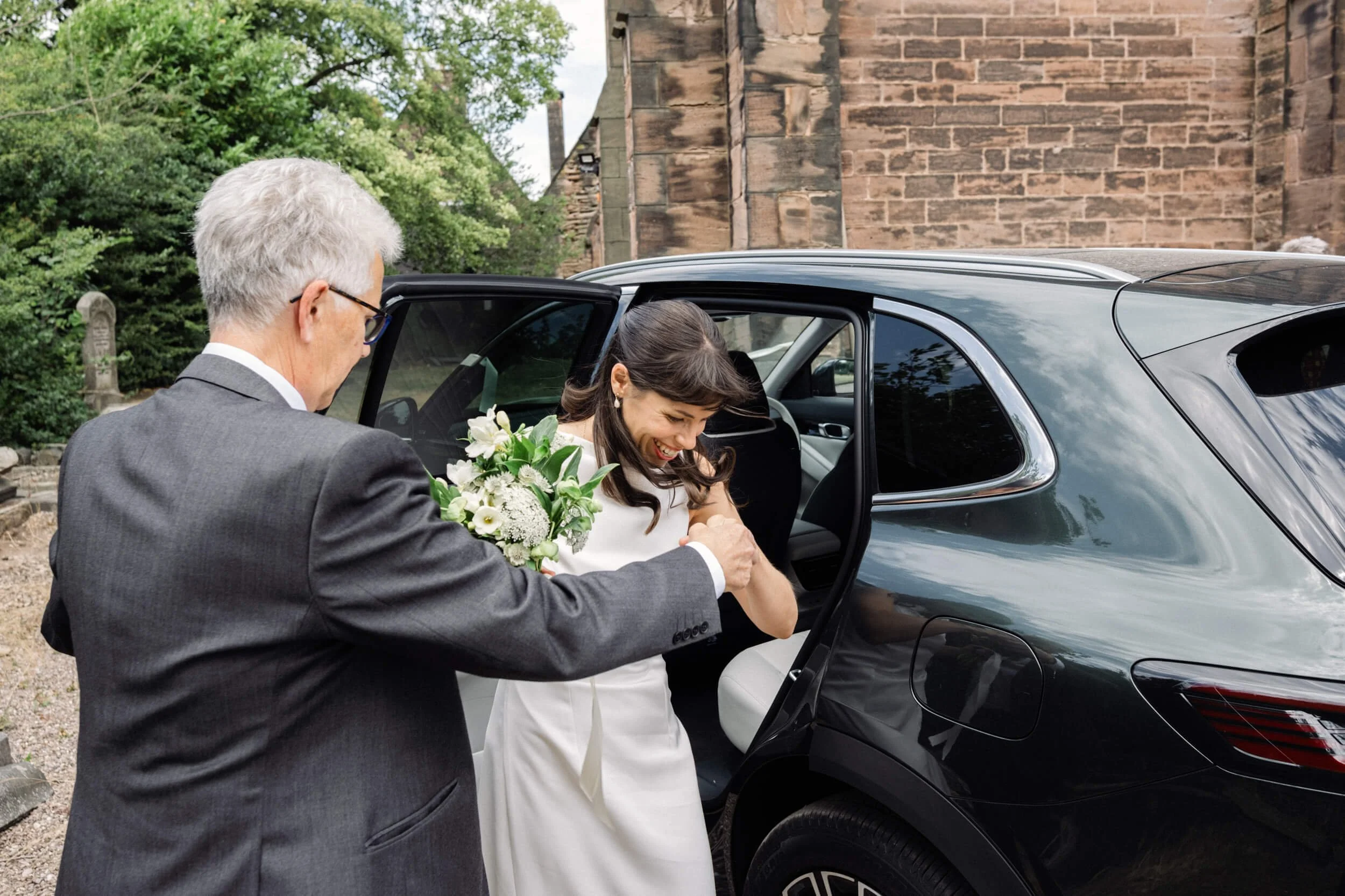 Bride steps out of the car outside the church assisted by her father.