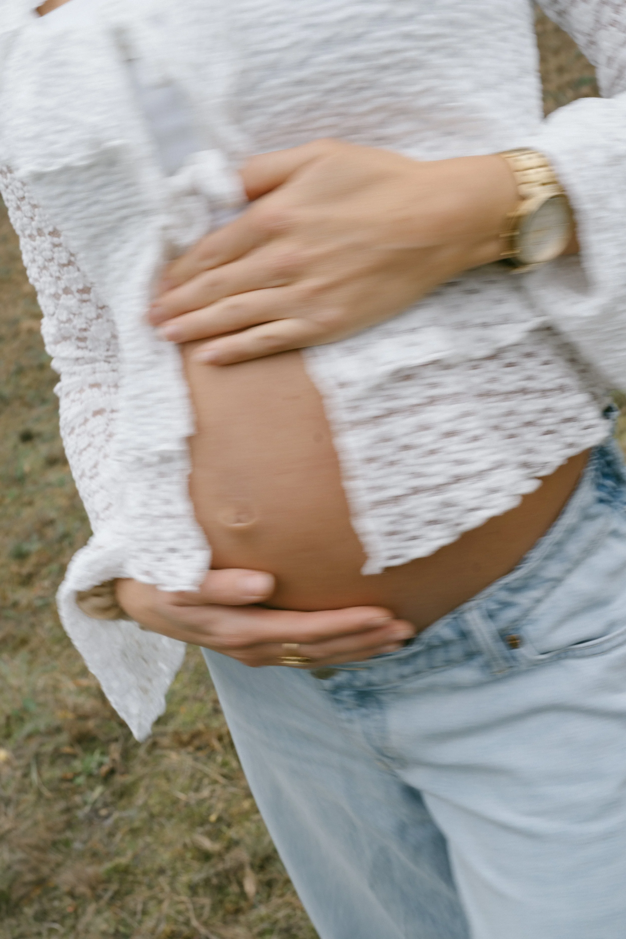 Close-up of a pregnant woman with a hand gently resting on her belly, wearing a white lace top and light blue jeans outdoor.