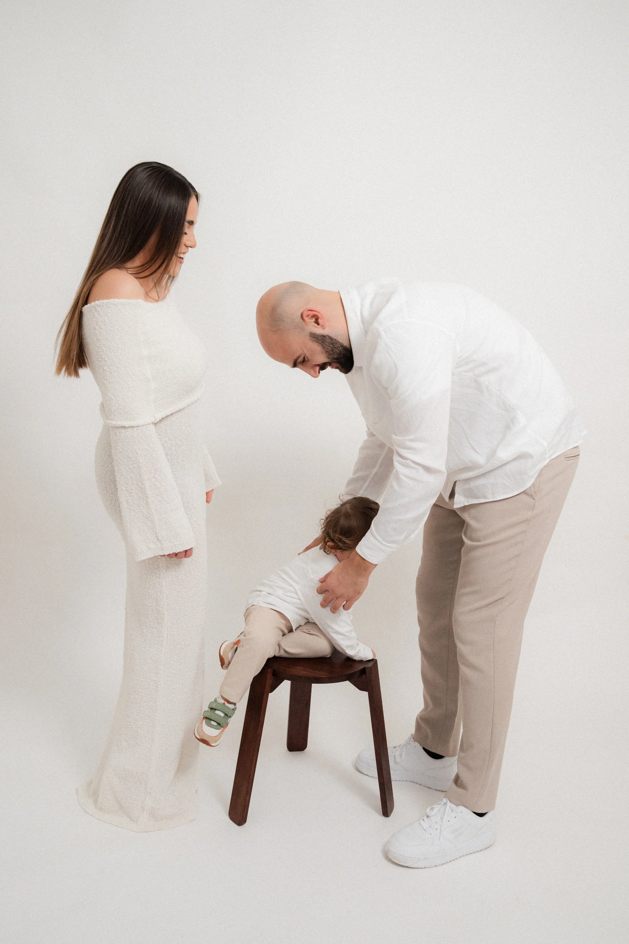 A family of three — a woman, a man, and a young child — interacting during a photoshoot in a white studio. The woman and man are smiling while the child is bent over a small wooden stool.