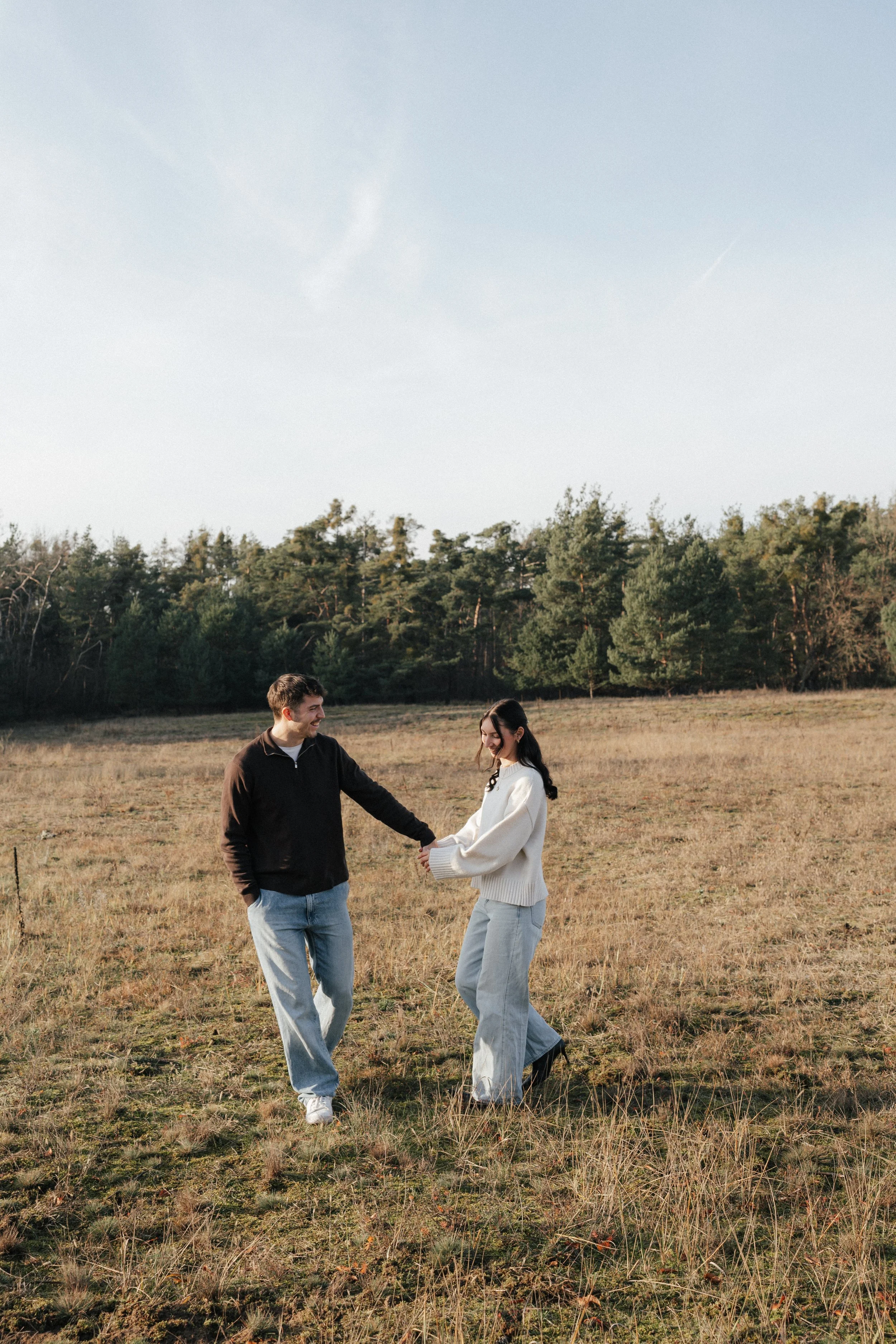 A couple holding hands and smiling in an open grassy field with trees in the background and a clear sky.