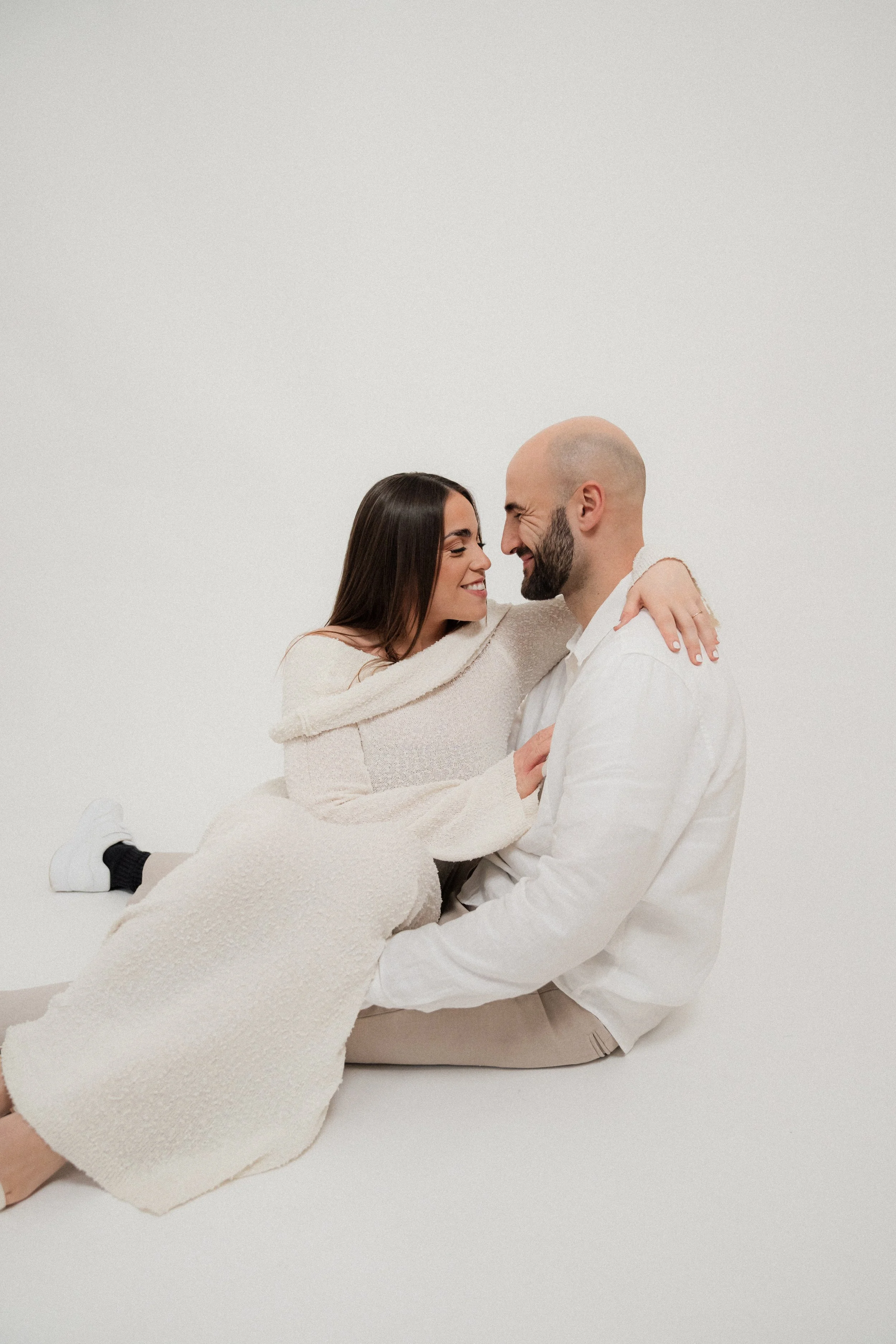 A couple sitting on the floor, facing each other, smiling and touching noses, with white background.
