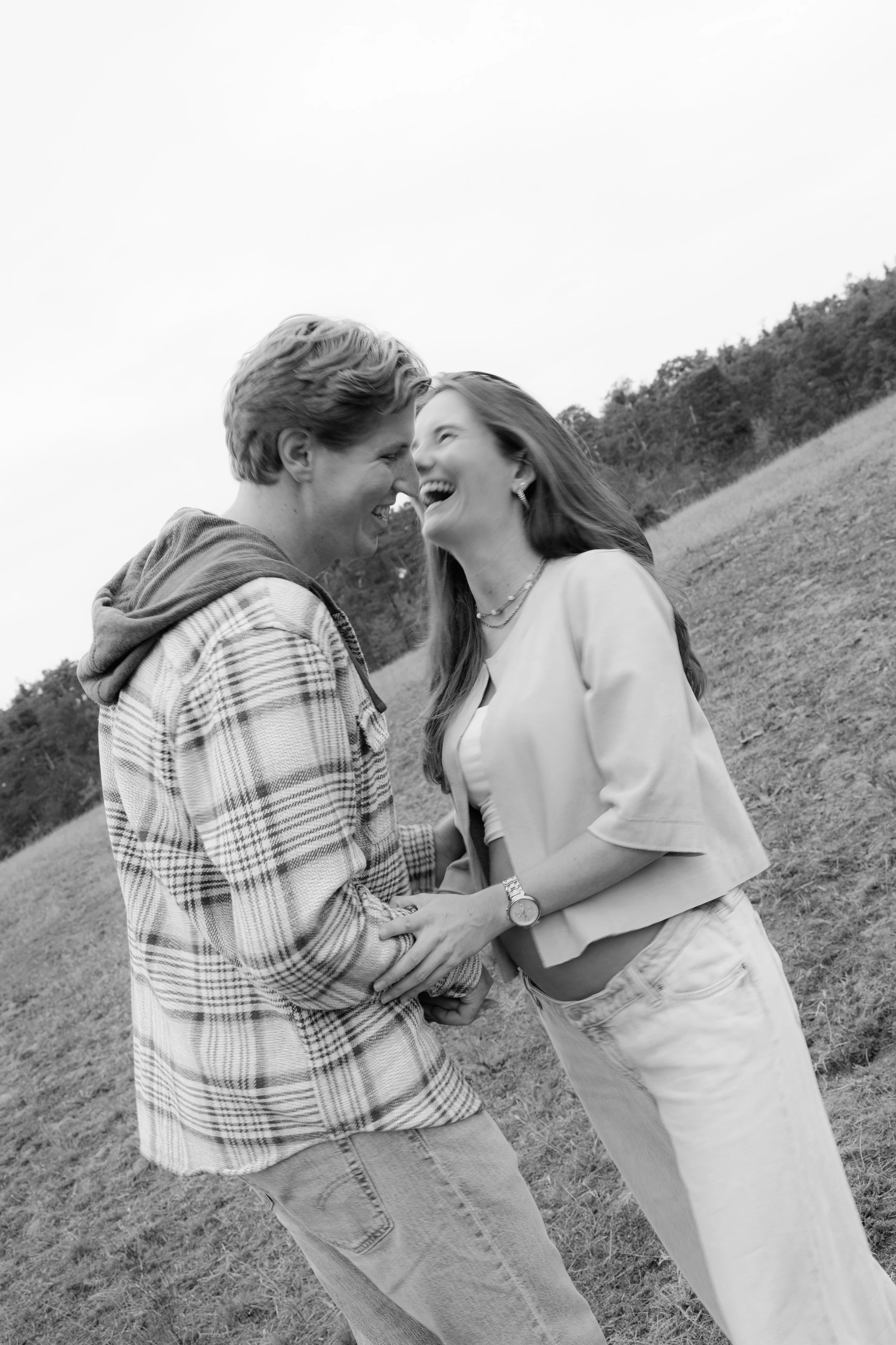 A black and white photo of a young couple laughing and holding hands outdoors in a grassy field.