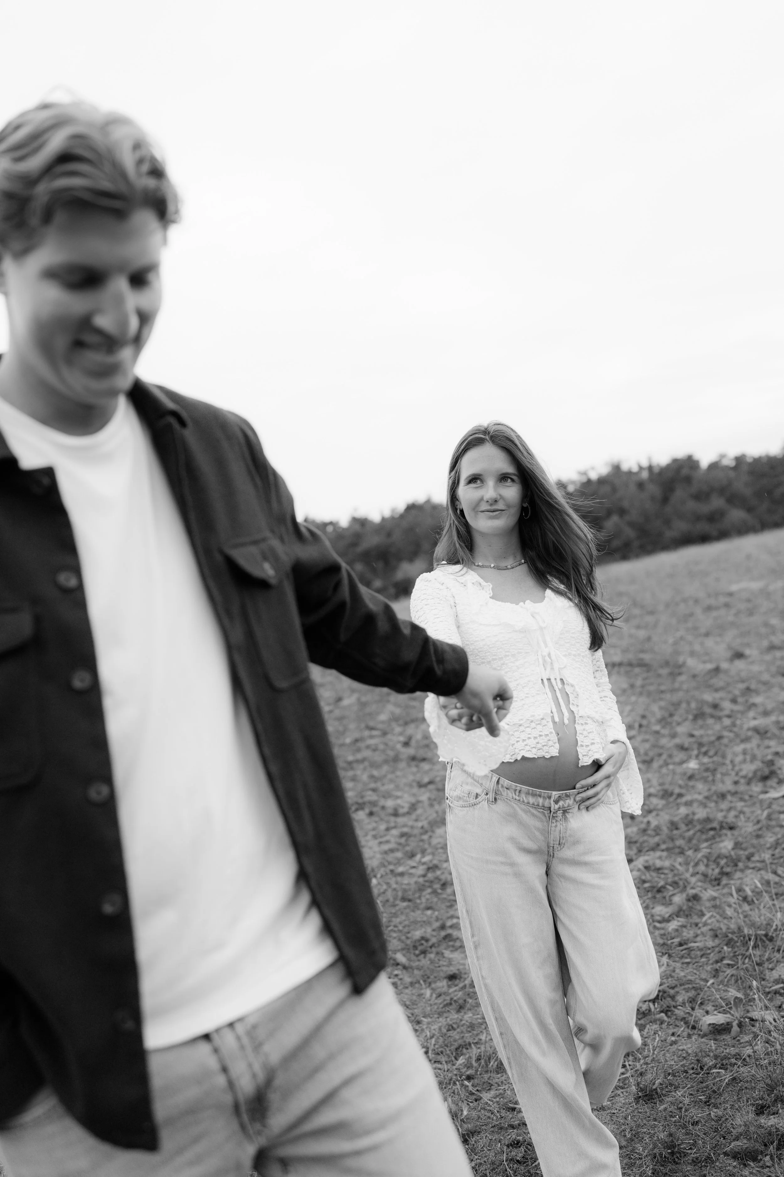 A black and white photo of a woman in a white top and baggy jeans holding her pregnant belly, being led by a man in a dark jacket and light shirt on a grassy field with trees in the background.
