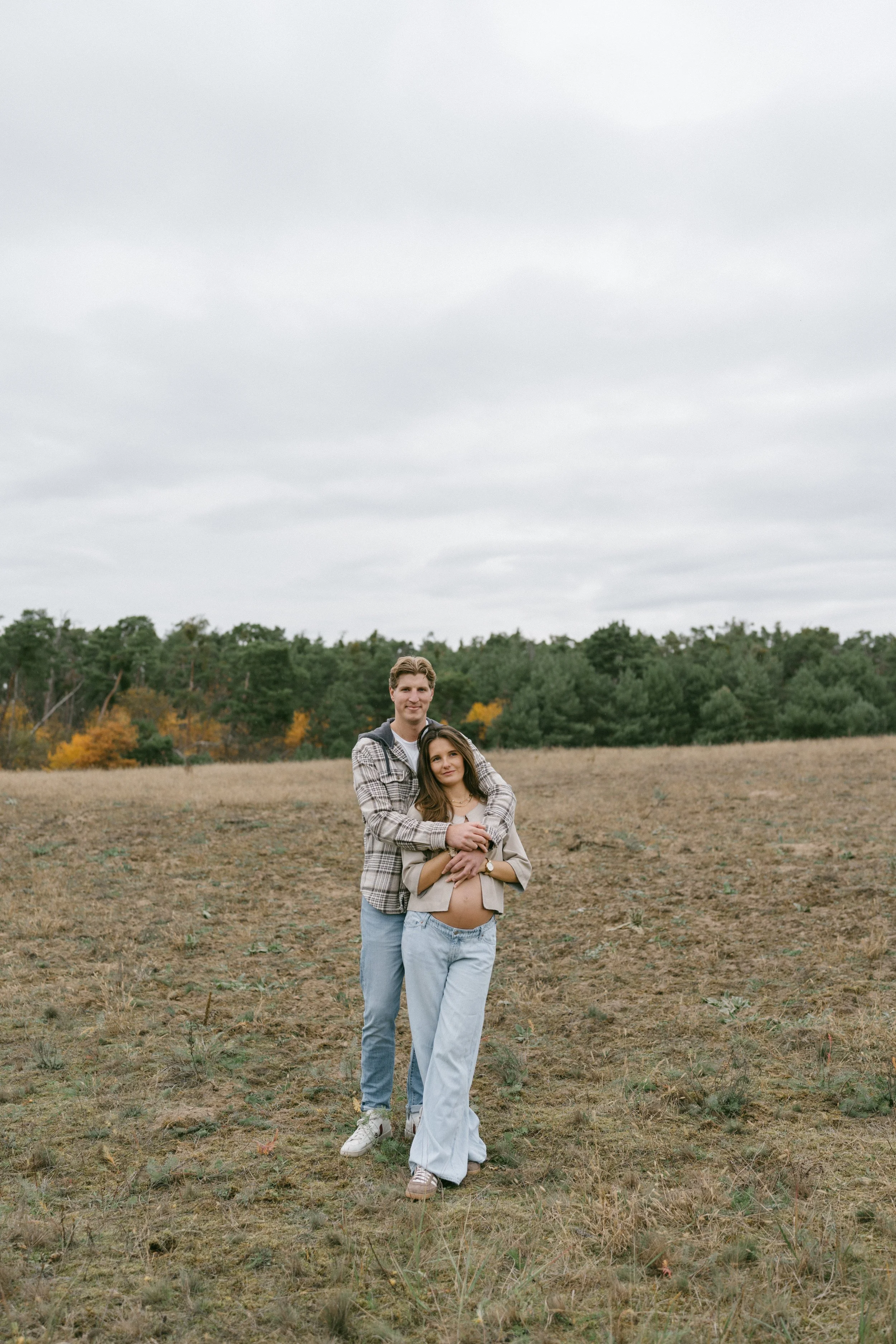 A couple outdoors in a field, with the man behind the woman, holding her and smiling, as the woman touches her pregnant belly.