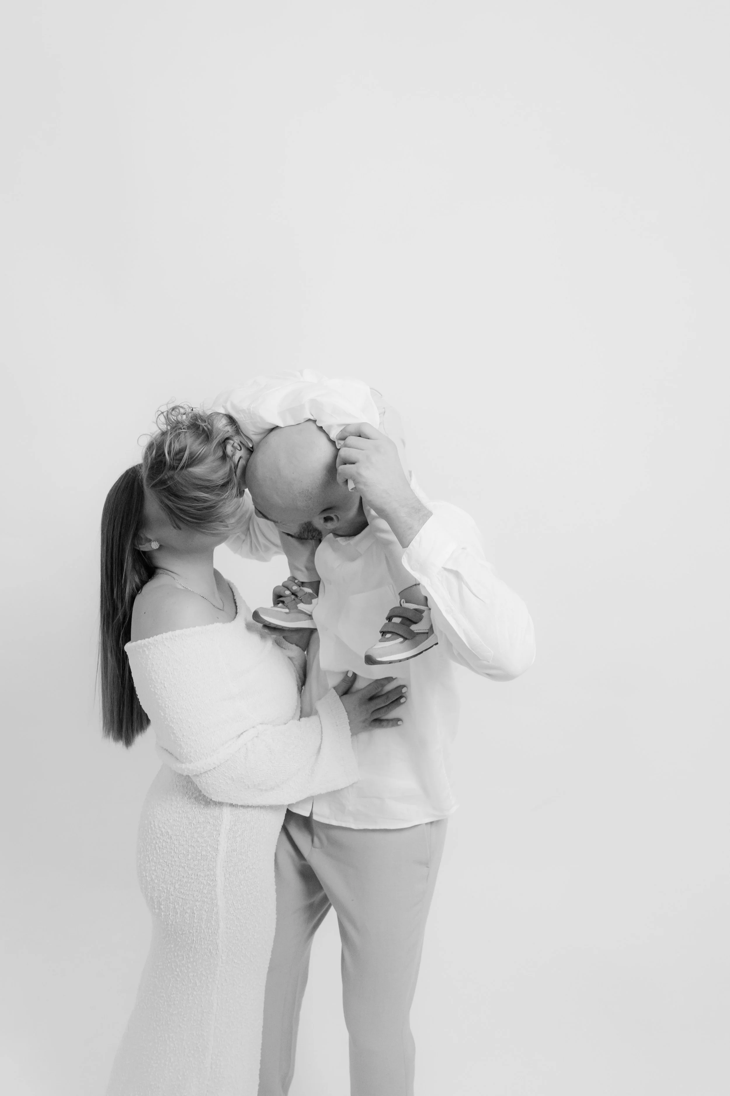 A couple holding their young child, giving each other a kiss, against a plain white background.