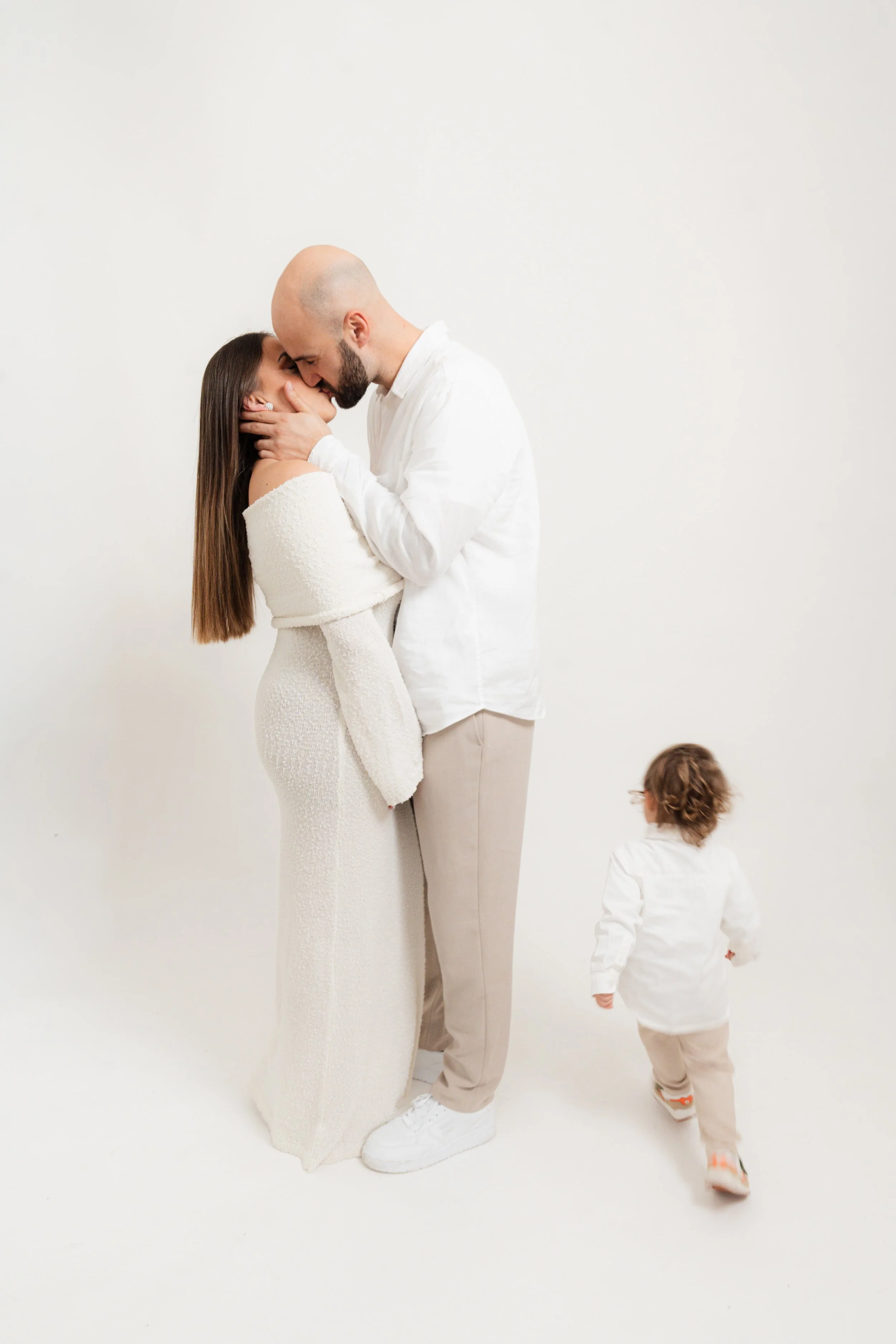 A couple sharing a kiss with a small child walking away in a white studio setting.