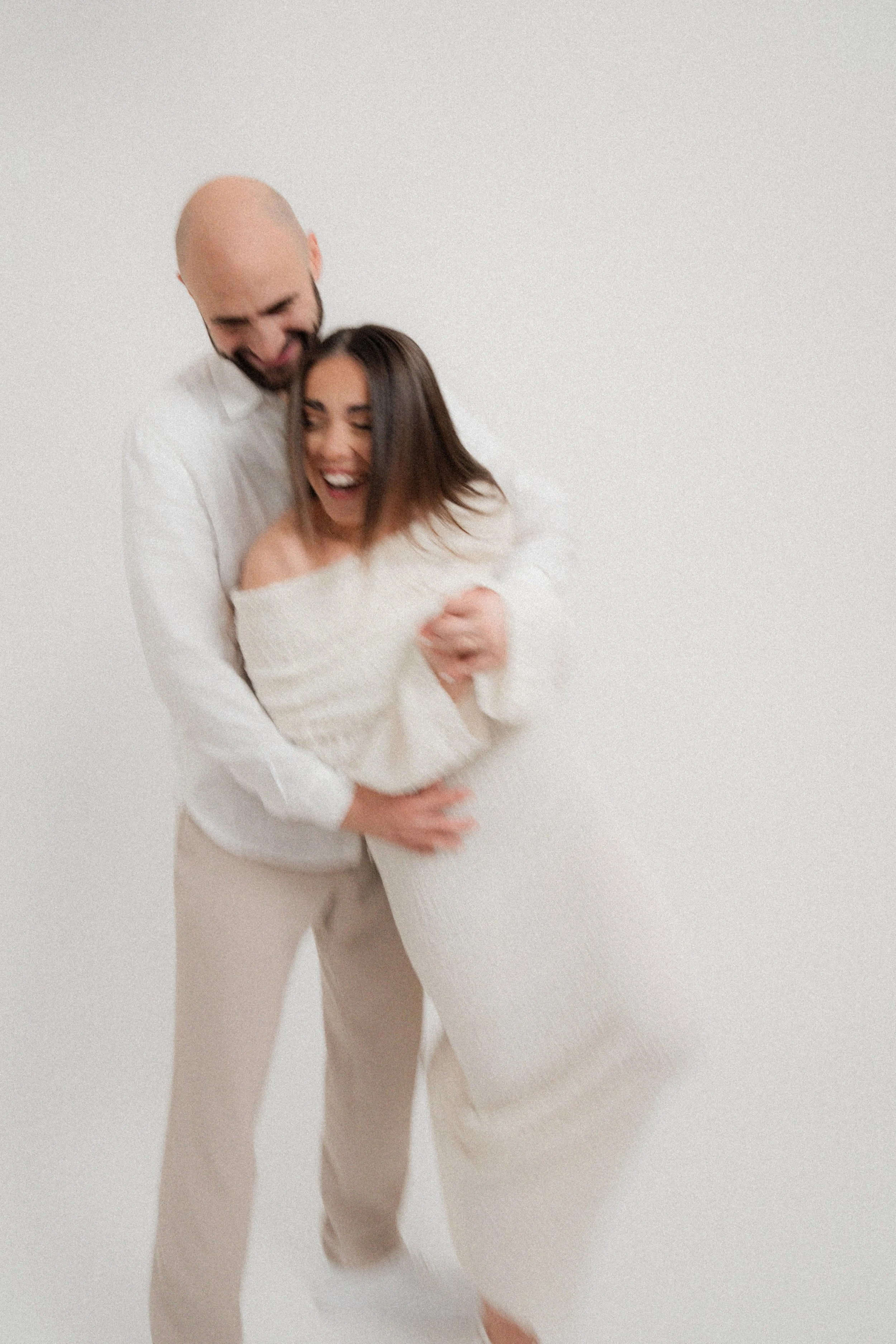 A man and woman laughing and hugging against a plain white background.