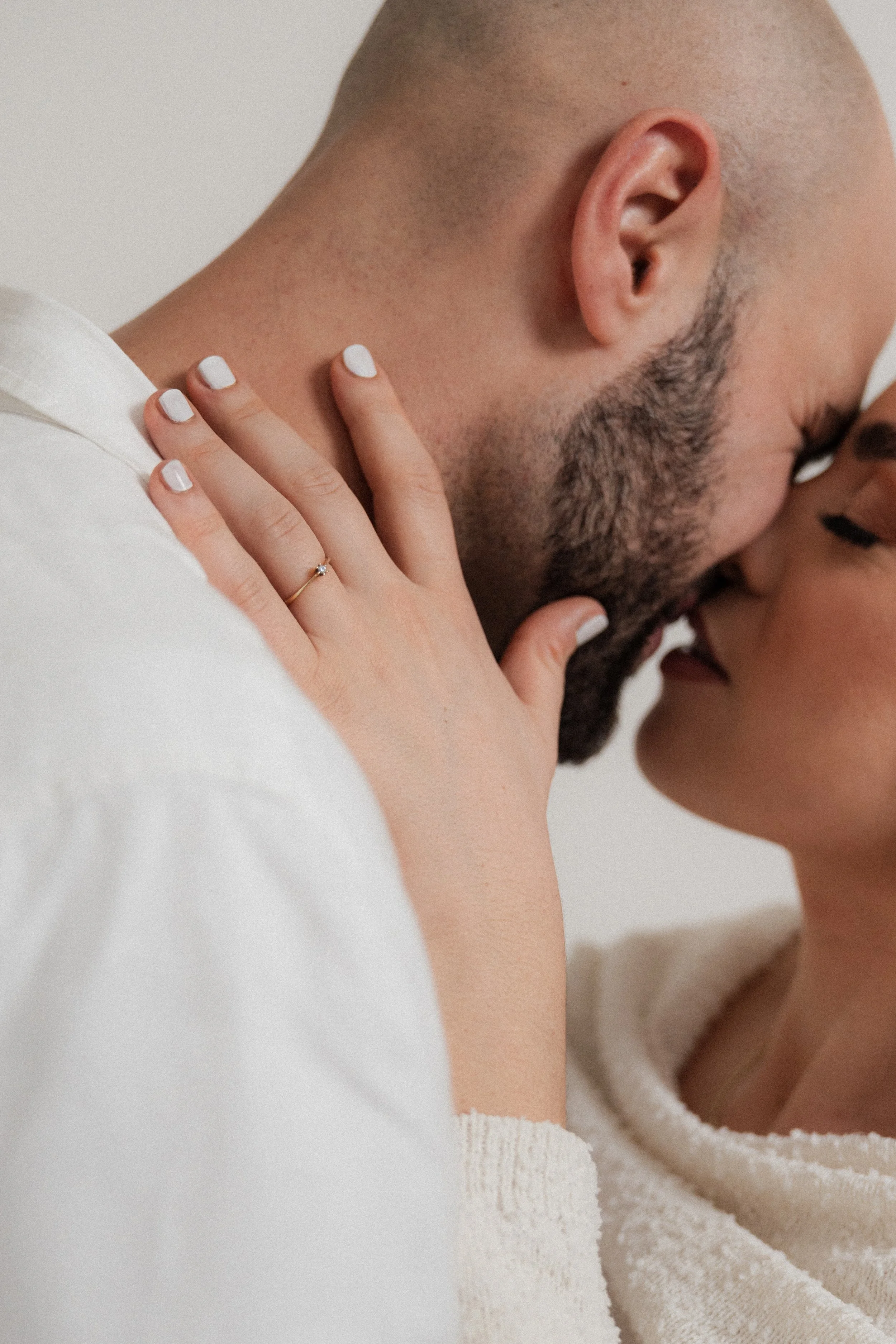 A close-up of a couple about to kiss, with the woman gently holding the man's face. The woman is wearing a ring on her finger, and both are wearing light-colored clothing.