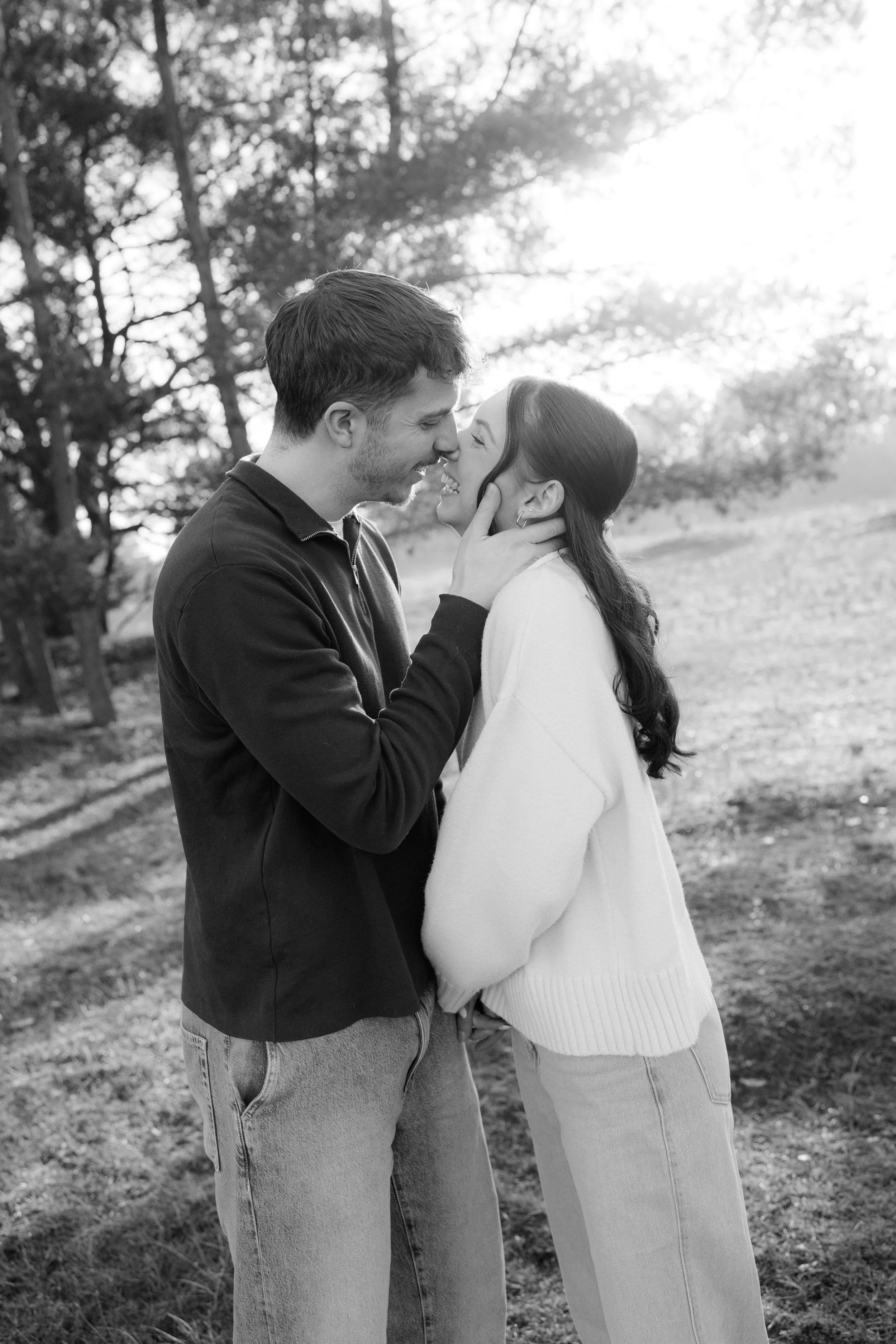 A black and white photo of a couple outdoors, smiling, with their faces close as they hold each other's faces, in a natural, wooded setting with sunlight filtering through the trees.