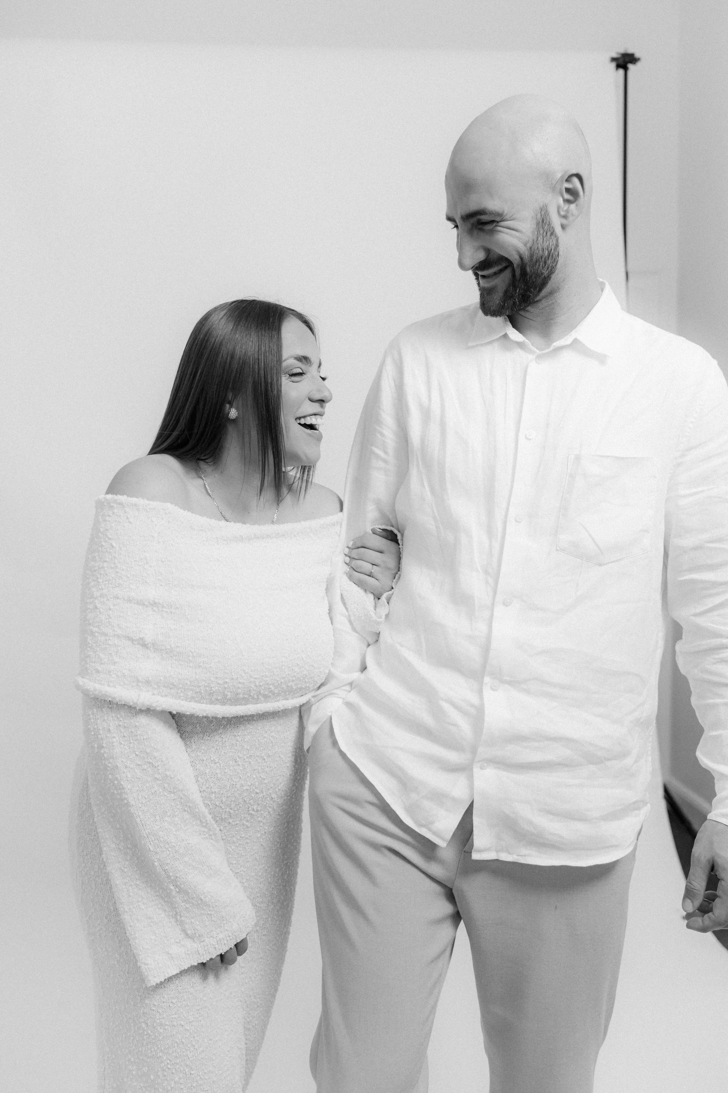 A woman and a man are smiling and laughing while holding hands in a studio setting. The woman has long hair and is wearing an off-the-shoulder sweater, while the man is bald, has a beard, and is dressed in a white button-up shirt.