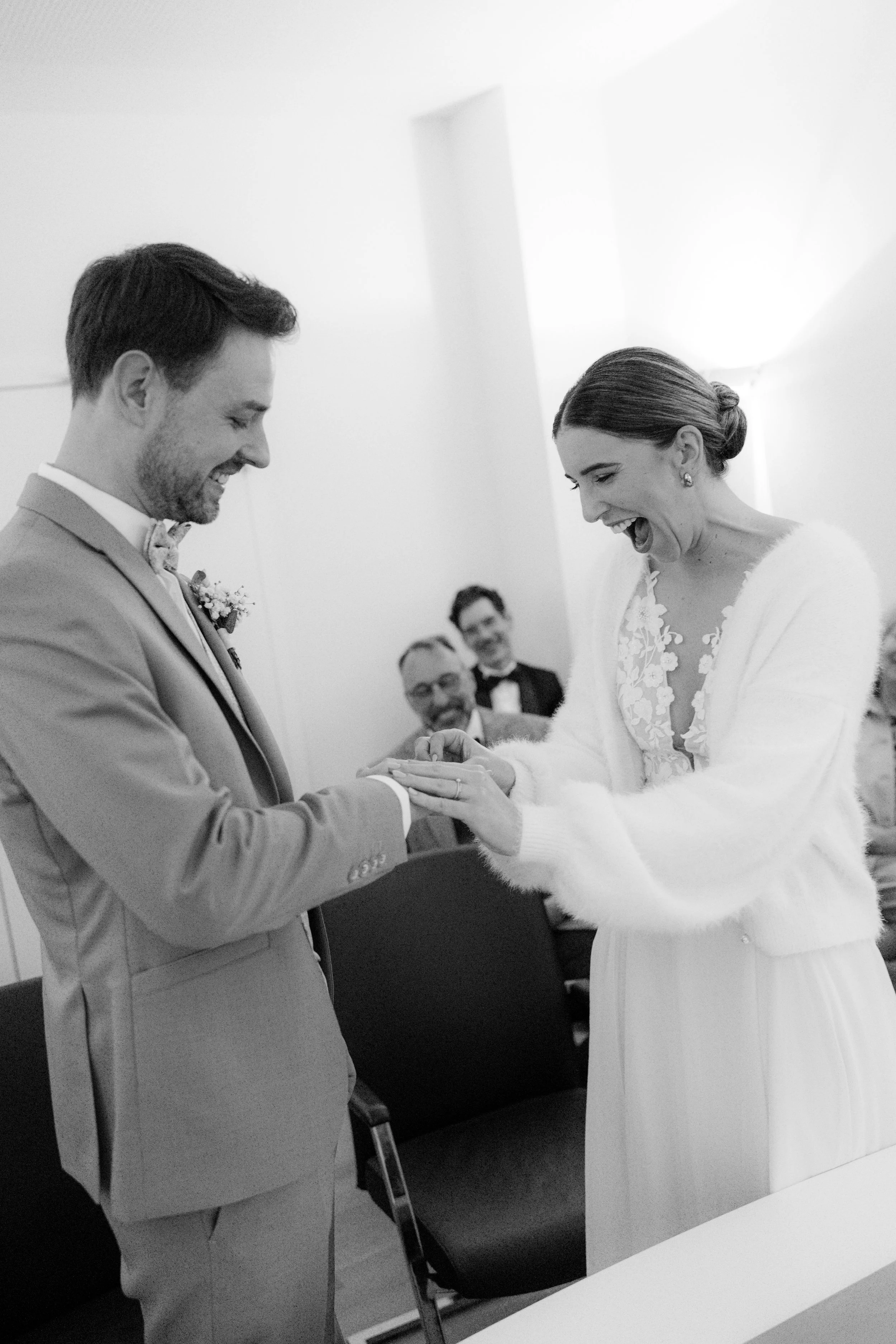 A black and white photo of a wedding ceremony, with a bride and groom exchanging rings and happy guests in the background.