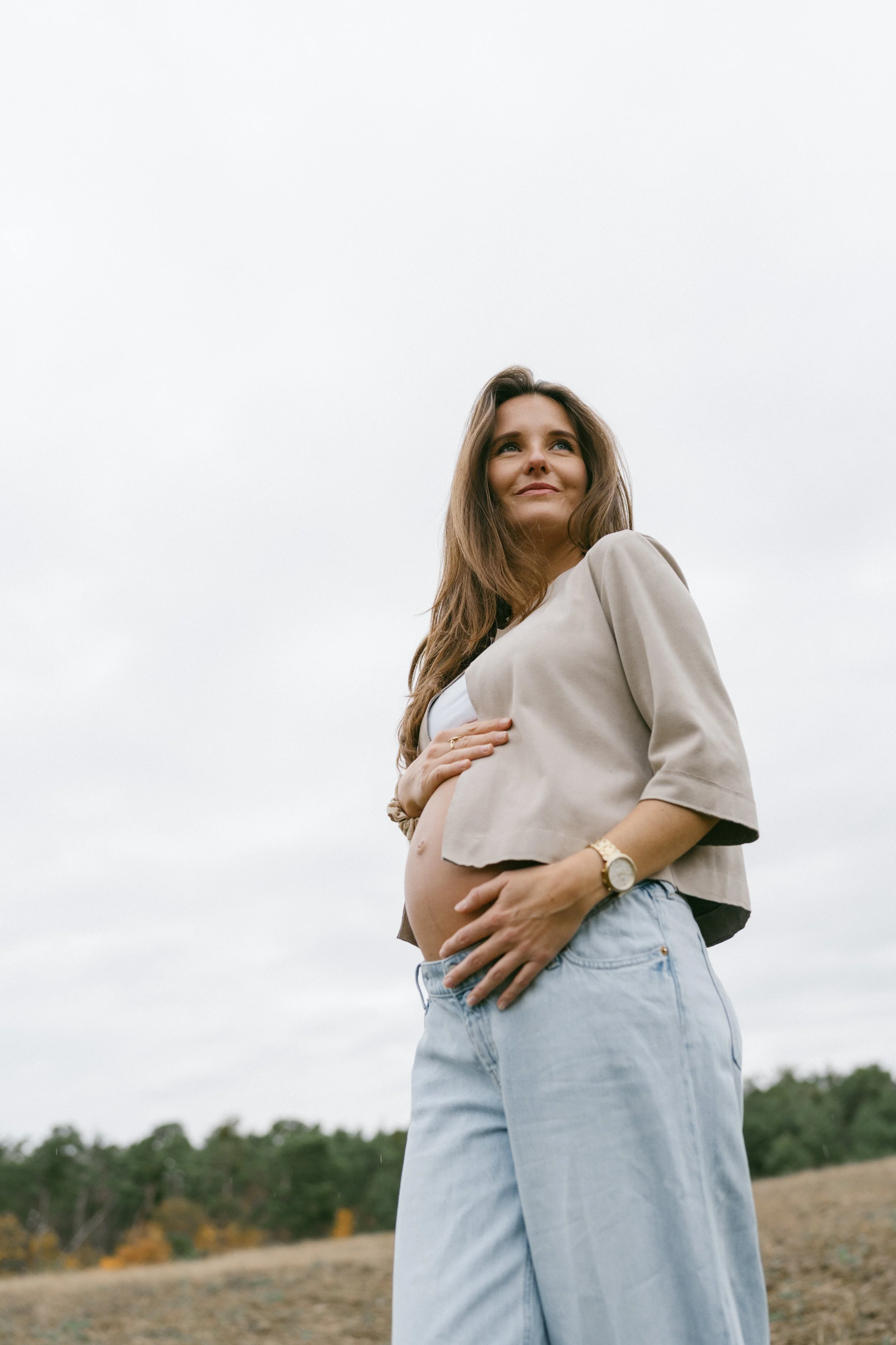 A pregnant woman standing outdoors on a cloudy day, holding her belly and looking up with a gentle smile.