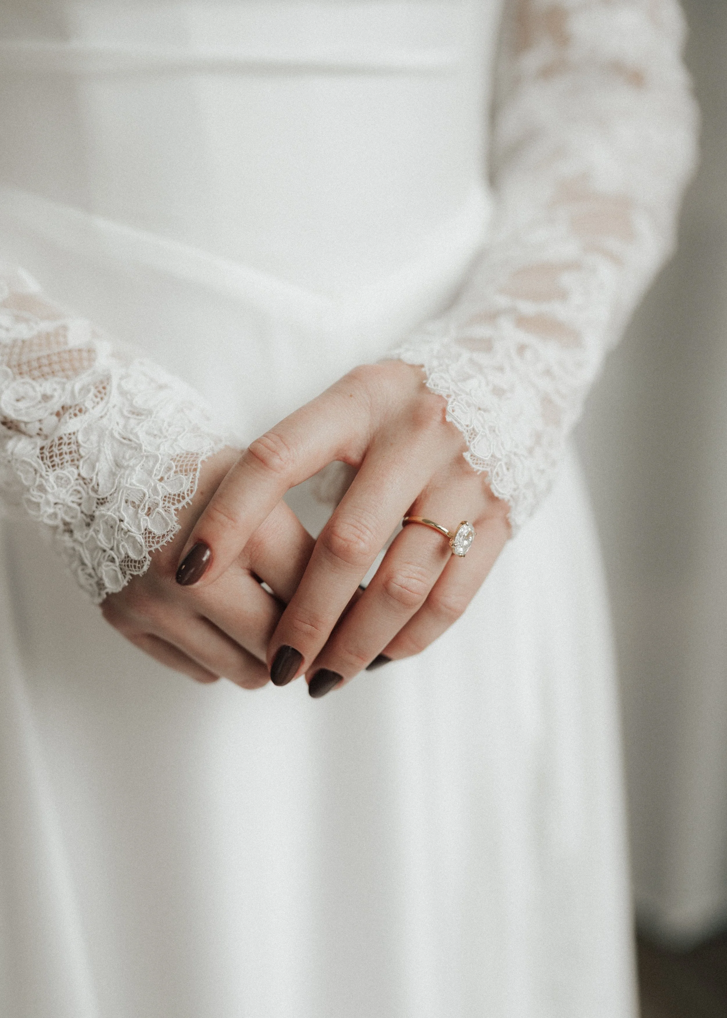 Close-up of a woman's hands clasped, wearing a white lace dress and a gold ring with a large gemstone.