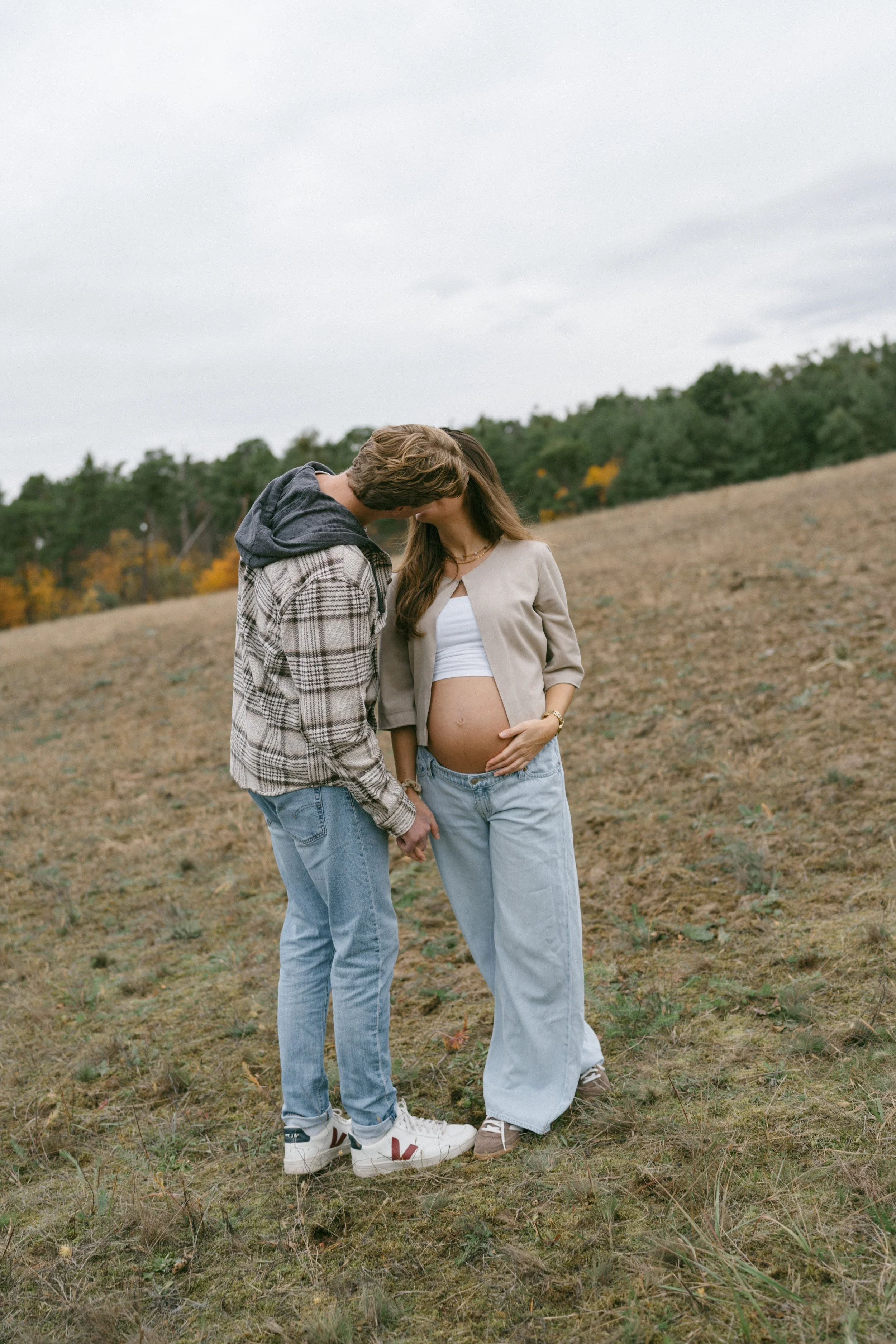 A pregnant woman and a man stand close together outdoors on a grassy field, sharing a kiss while holding hands. The woman is wearing a beige jacket, white crop top, and loose jeans, with her hand resting on her baby bump. The man is dressed in a plai