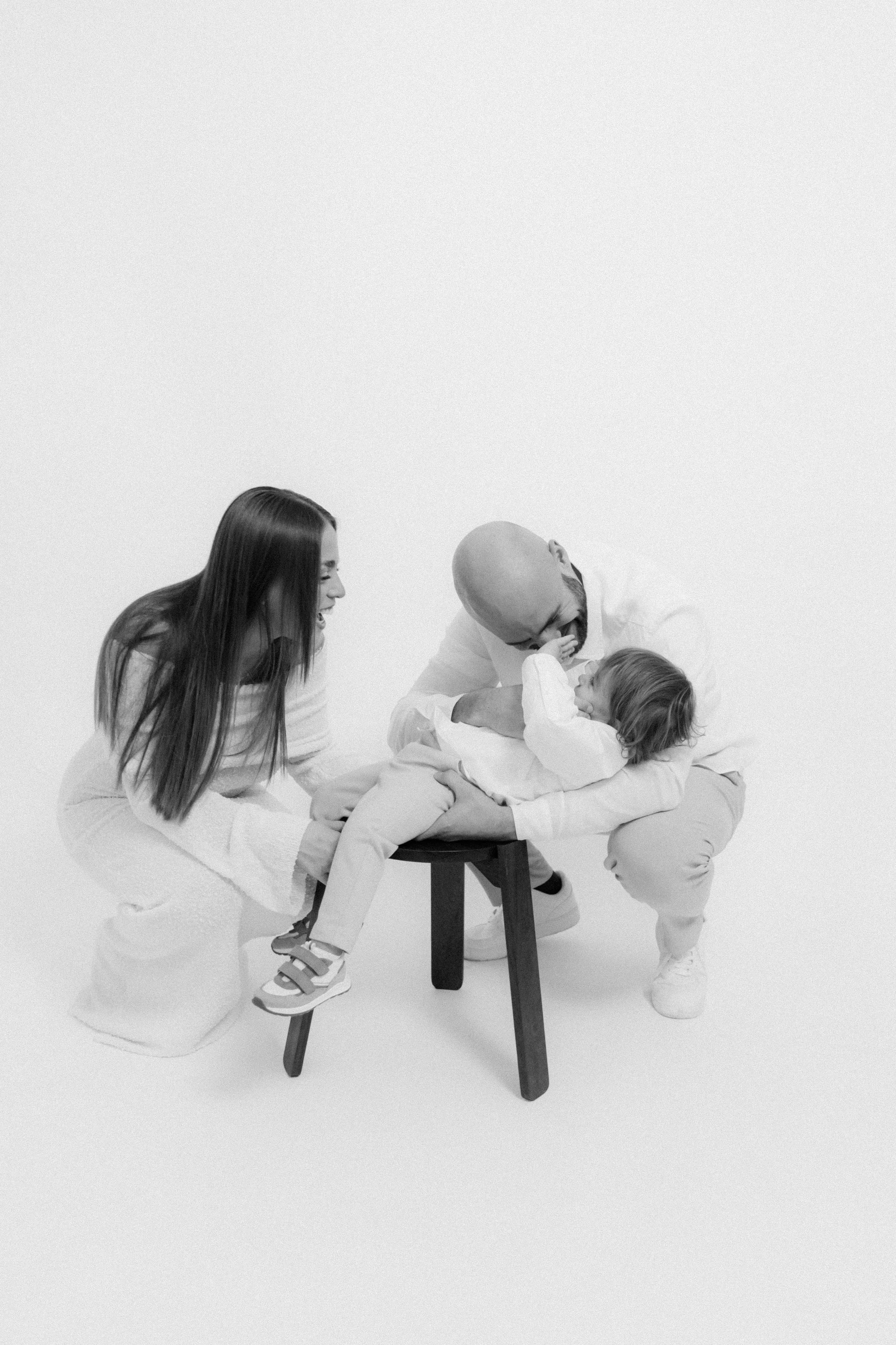 Black and white photo of a family with a father, mother, and young child on a small stool, laughing and playing together.
