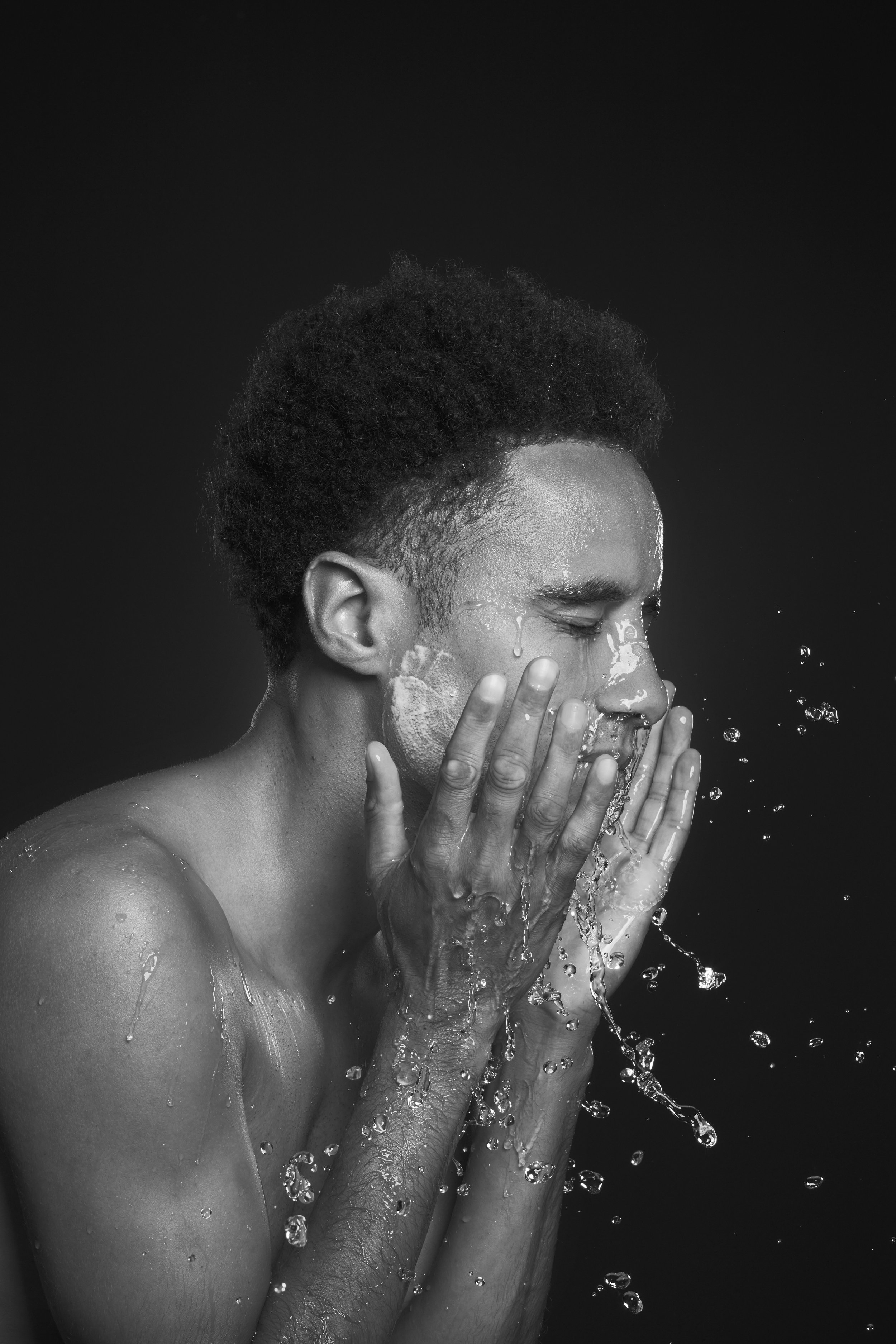 Black and white image of a person washing their face with water, hands cupping water, droplets splashing, against a dark background.
