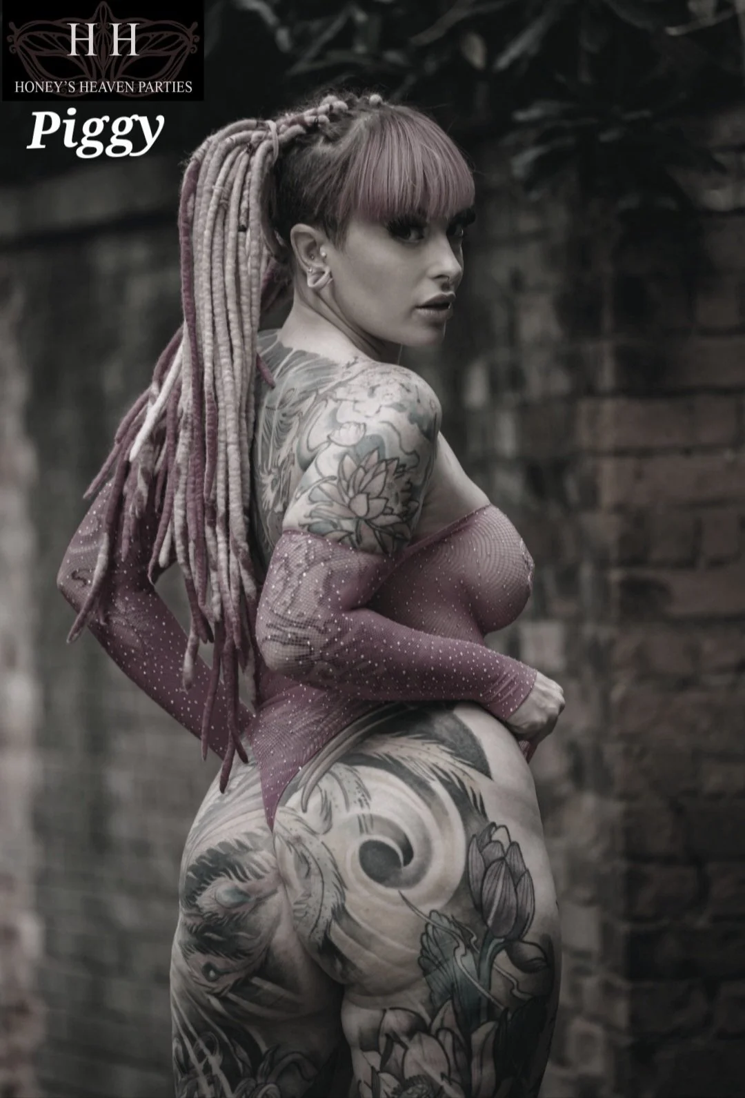 A tattooed woman with pink dreadlocks and bangs, wearing sheer, off-the-shoulder fabric, stands outdoors against a brick wall, looking over her shoulder. at Honey's Heaven Parties and Exclusive Elite Adult Event Mayfair London.