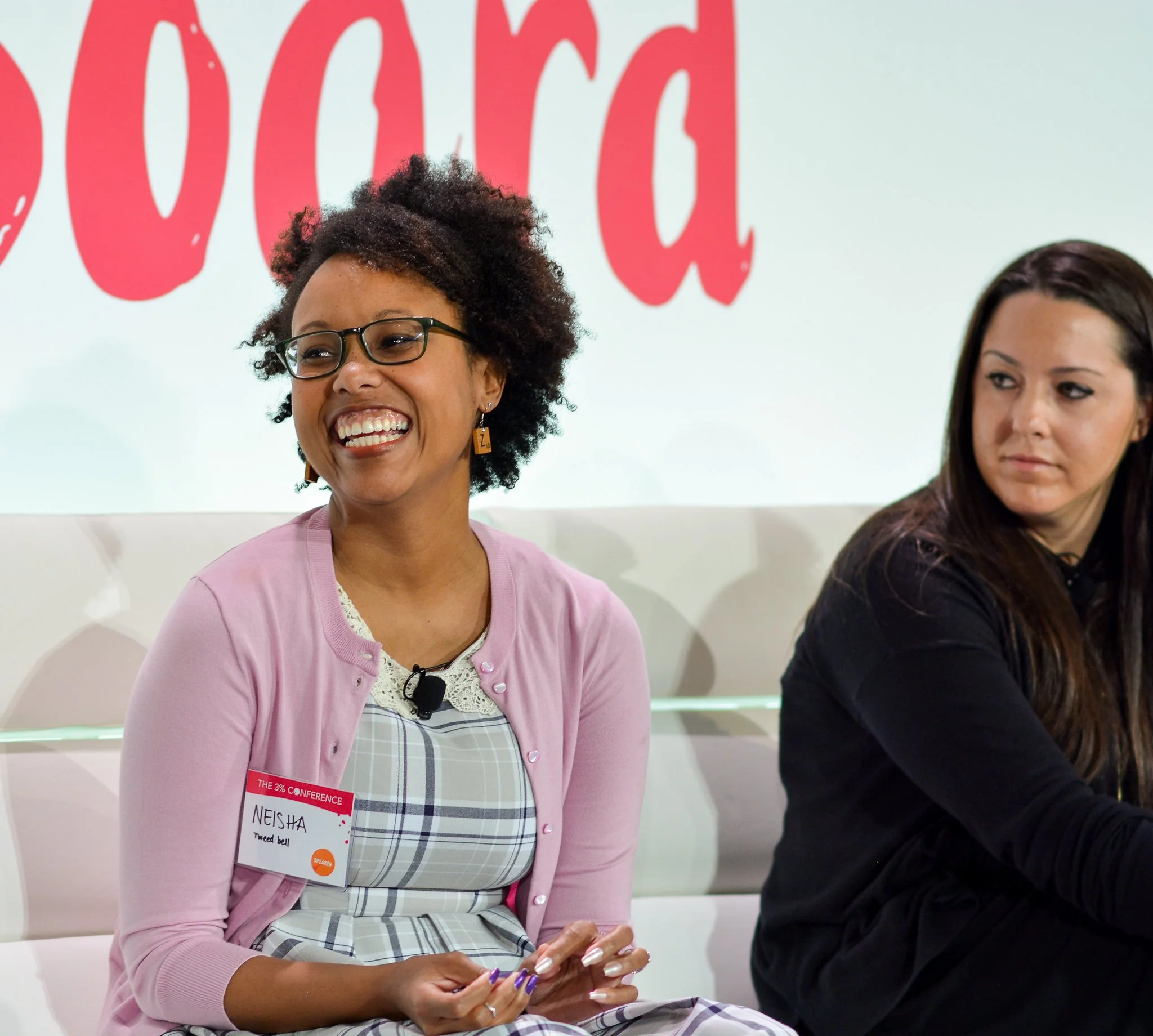Smiling woman with curly hair and glasses wearing a pink cardigan and plaid dress at conference, sitting beside a woman with long dark hair, in front of red letters on a white wall.