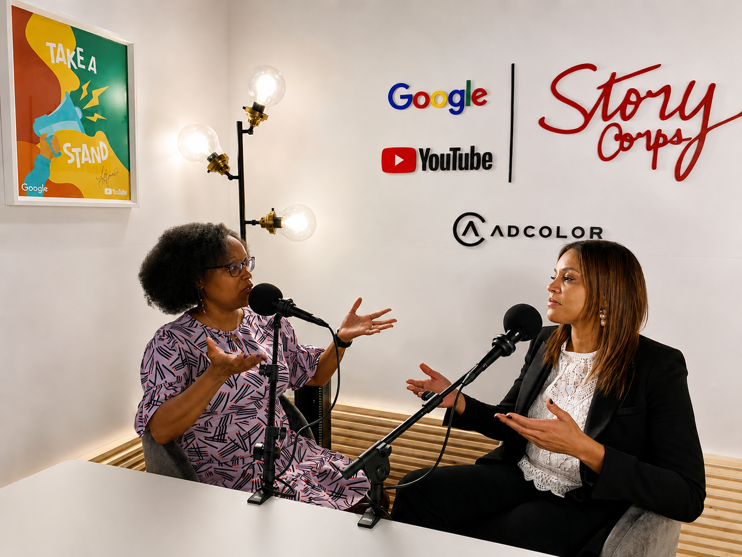 Two women are sitting at a table in a podcast studio, speaking into microphones while engaging in conversation. The woman on the left has curly hair, wears glasses, and a colorful dress, gesturing with her hands. The woman on the right has straight hair, wears a black blazer and white lace top, also gesturing with her hands. The background features wall decor with logos of Google, YouTube, ADColor, and the phrase 'Story Corps' in red lettering. A framed poster on the wall says 'TAKE A STAND' with an illustration of a raised fist.