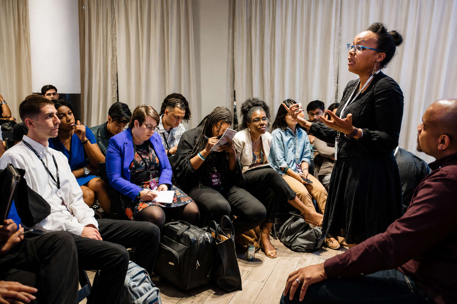 A diverse group of people attending a presentation or workshop, sitting attentively and taking notes, with a woman standing and speaking in front of them.