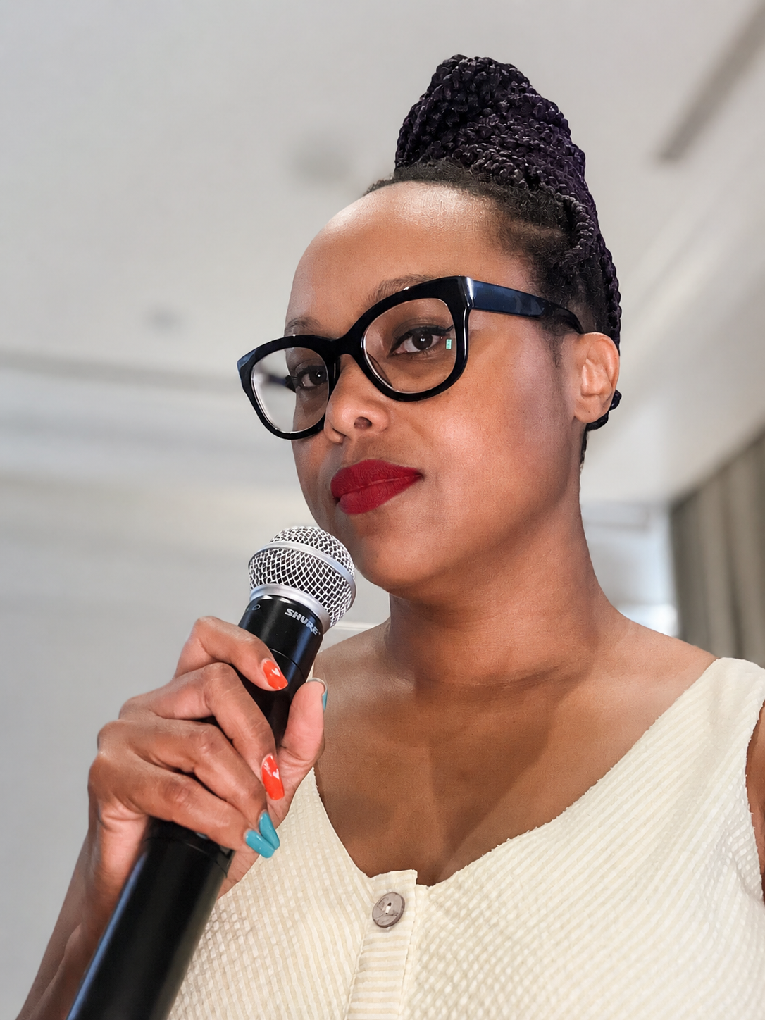 A woman with dark skin, wearing large black glasses, holding a microphone, with red lipstick and multicolored nails.