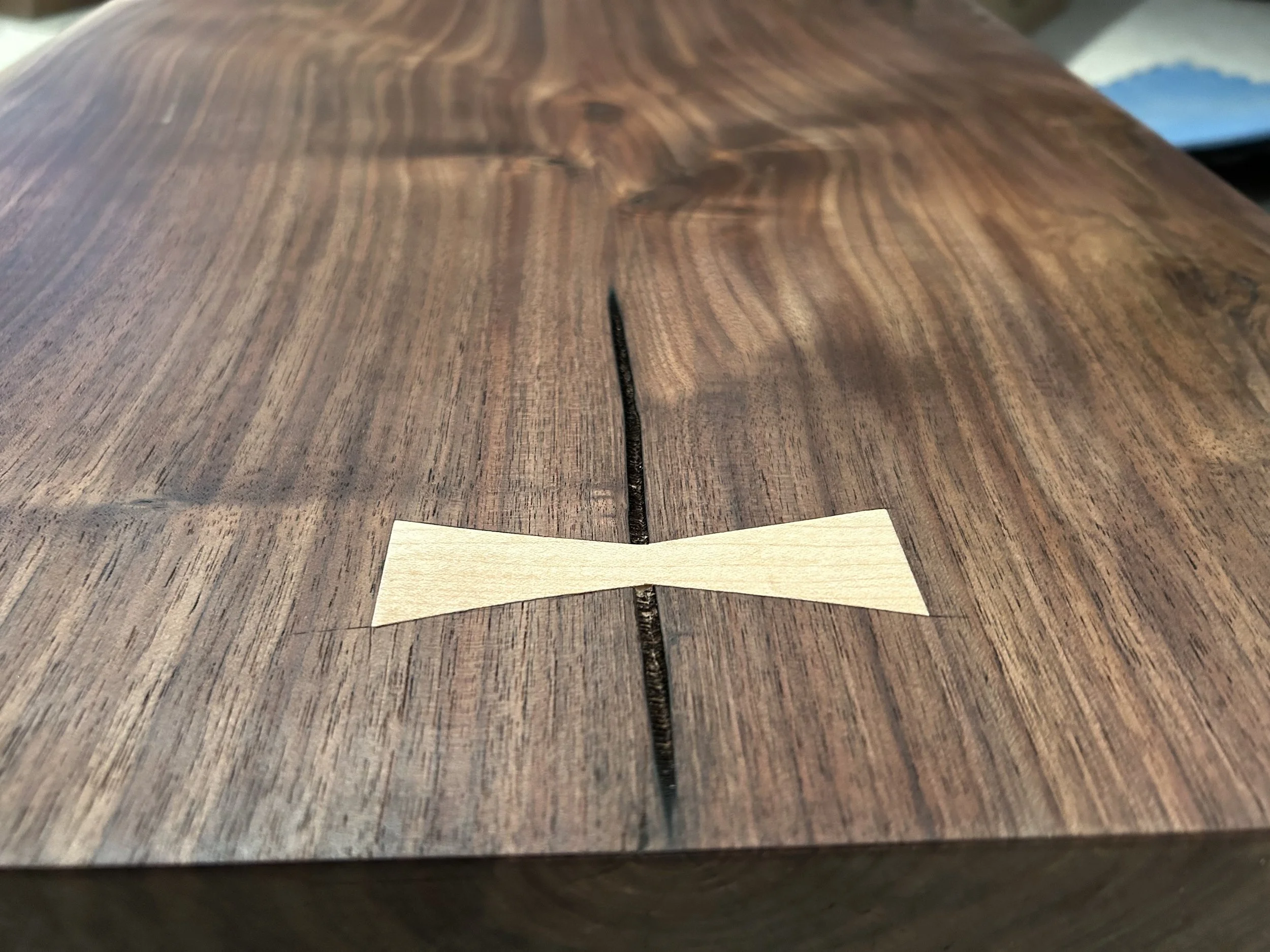 Close-up view of a wooden surface with a small, light-colored wooden bowtie-shaped inlay in the center, and a split running along the grain.