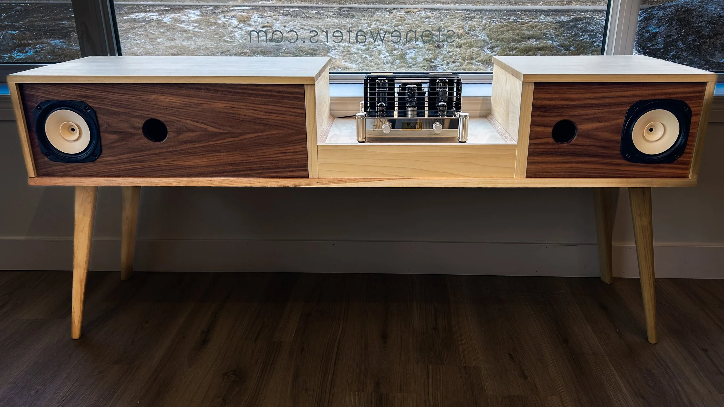 A wooden sideboard with two speaker boxes and a vacuum tube amplifier on top, placed in front of a window.