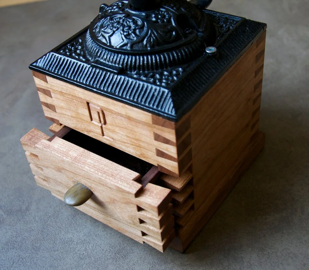 A vintage wooden coffee grinder with a carved black cast iron top and a small wooden drawer with a round brass knob.
