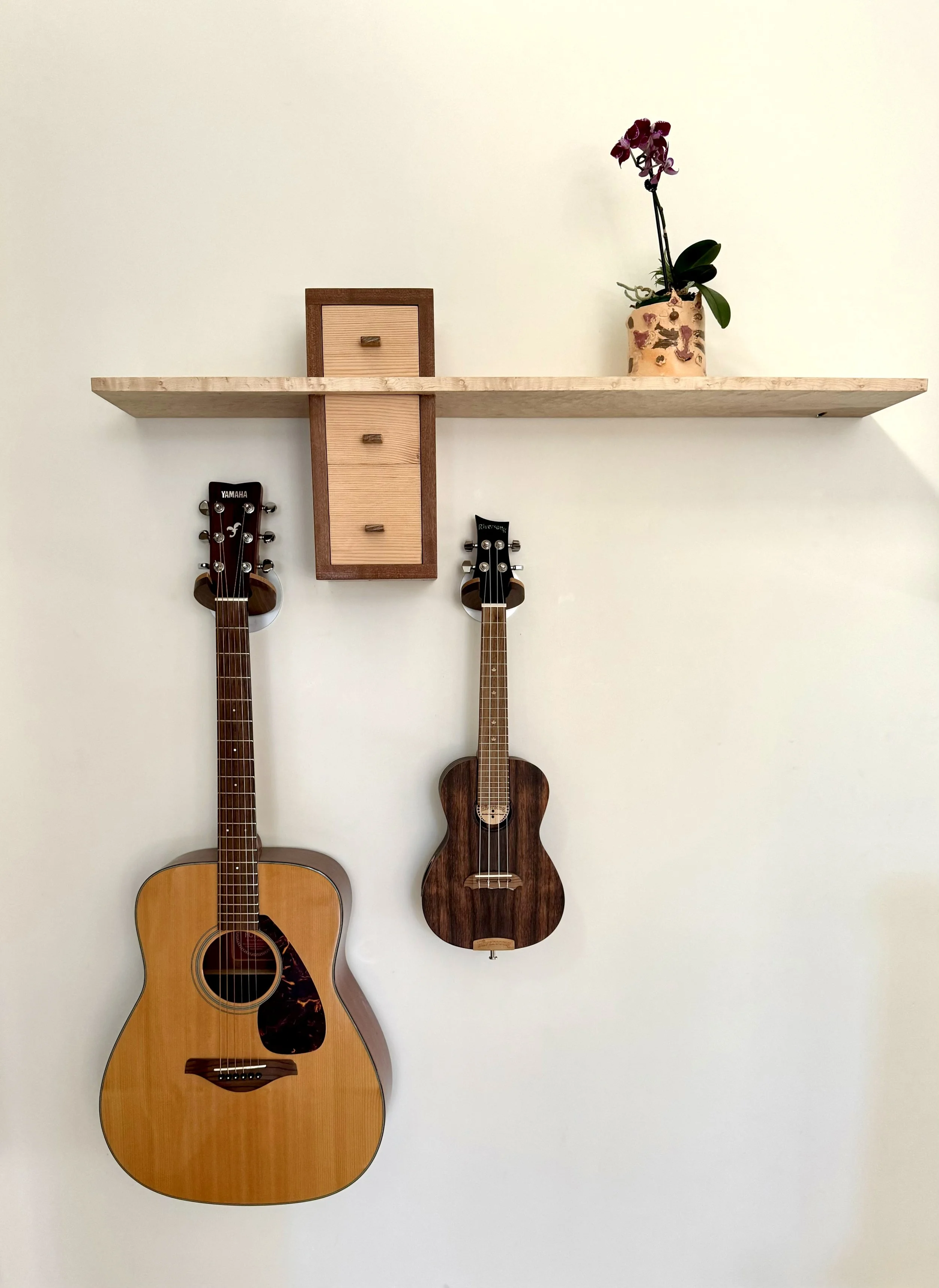 A wall decorated with two hanging guitars, a wooden wall-mounted cabinet, and a flowerpot with a purple orchid on a wooden shelf.