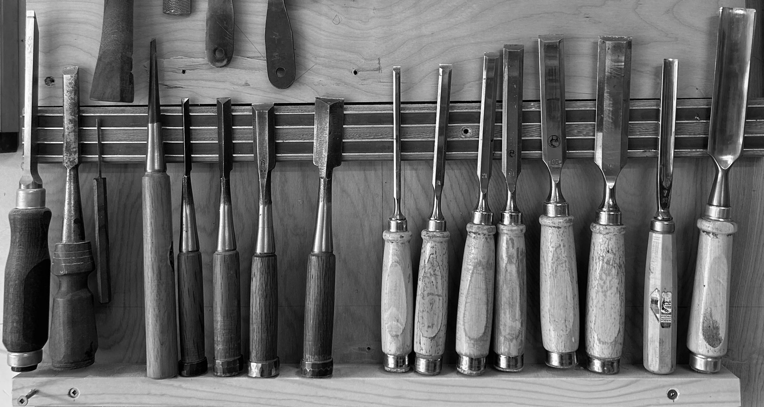 A row of woodworking chisels with wooden handles arranged upright on a wooden workbench with a pegboard holding some large chisels or hand tools in the background.