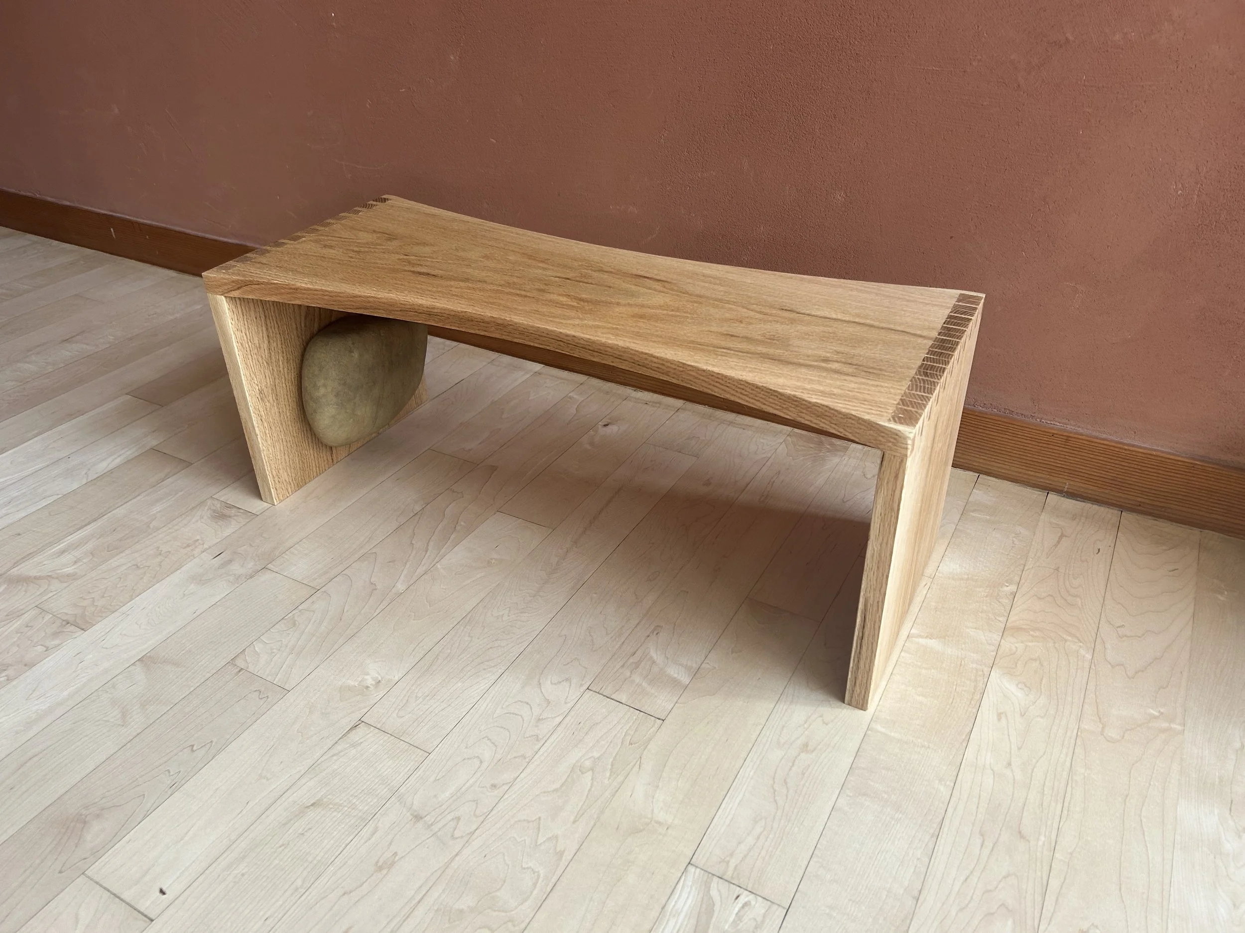 A wooden bench with a smooth stone embedded on one side, placed on a light-colored wooden floor against a reddish wall.