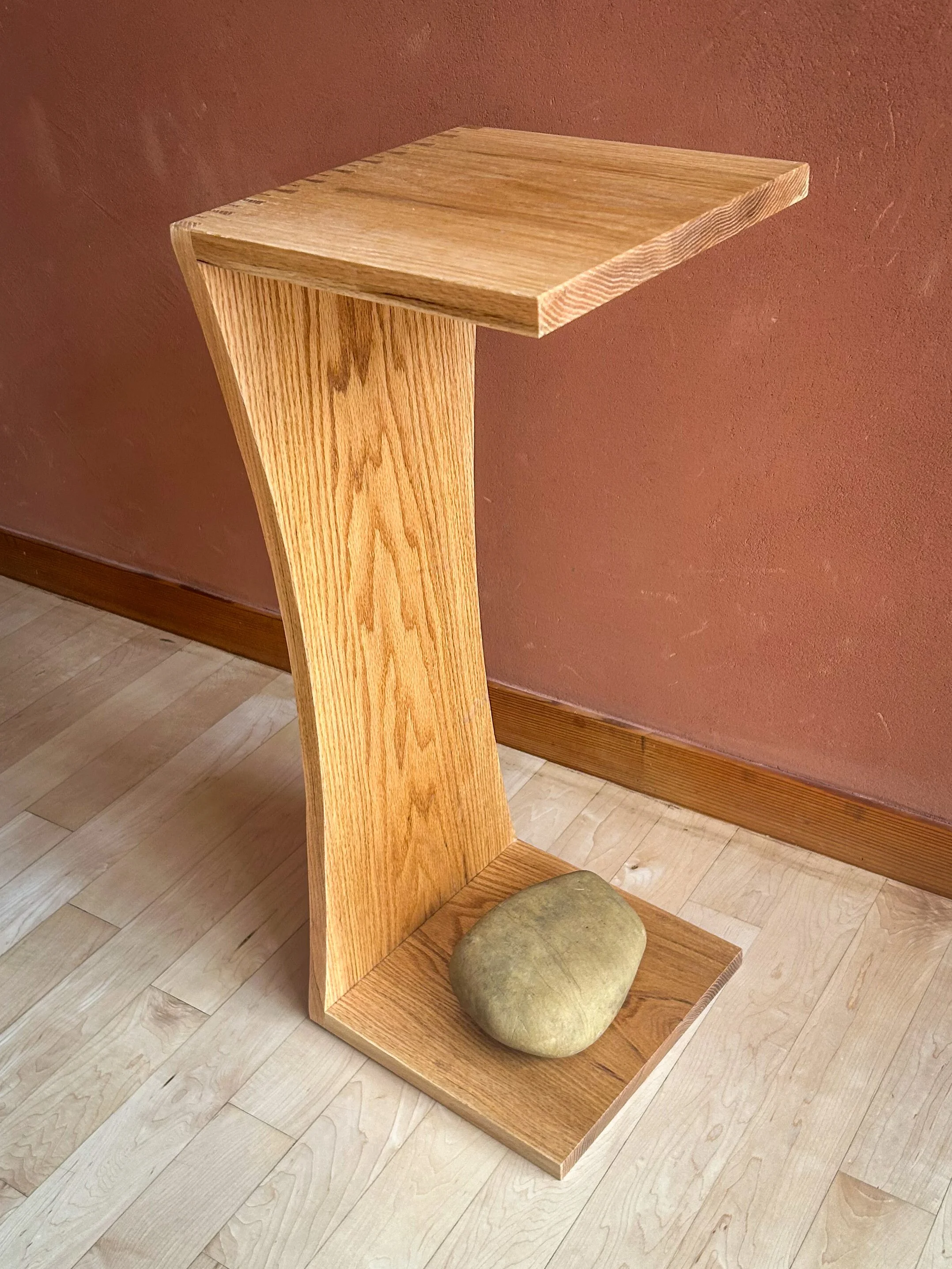 A wooden table with an angular support leg resting on a wooden floor, with a large stone placed on the lower platform.