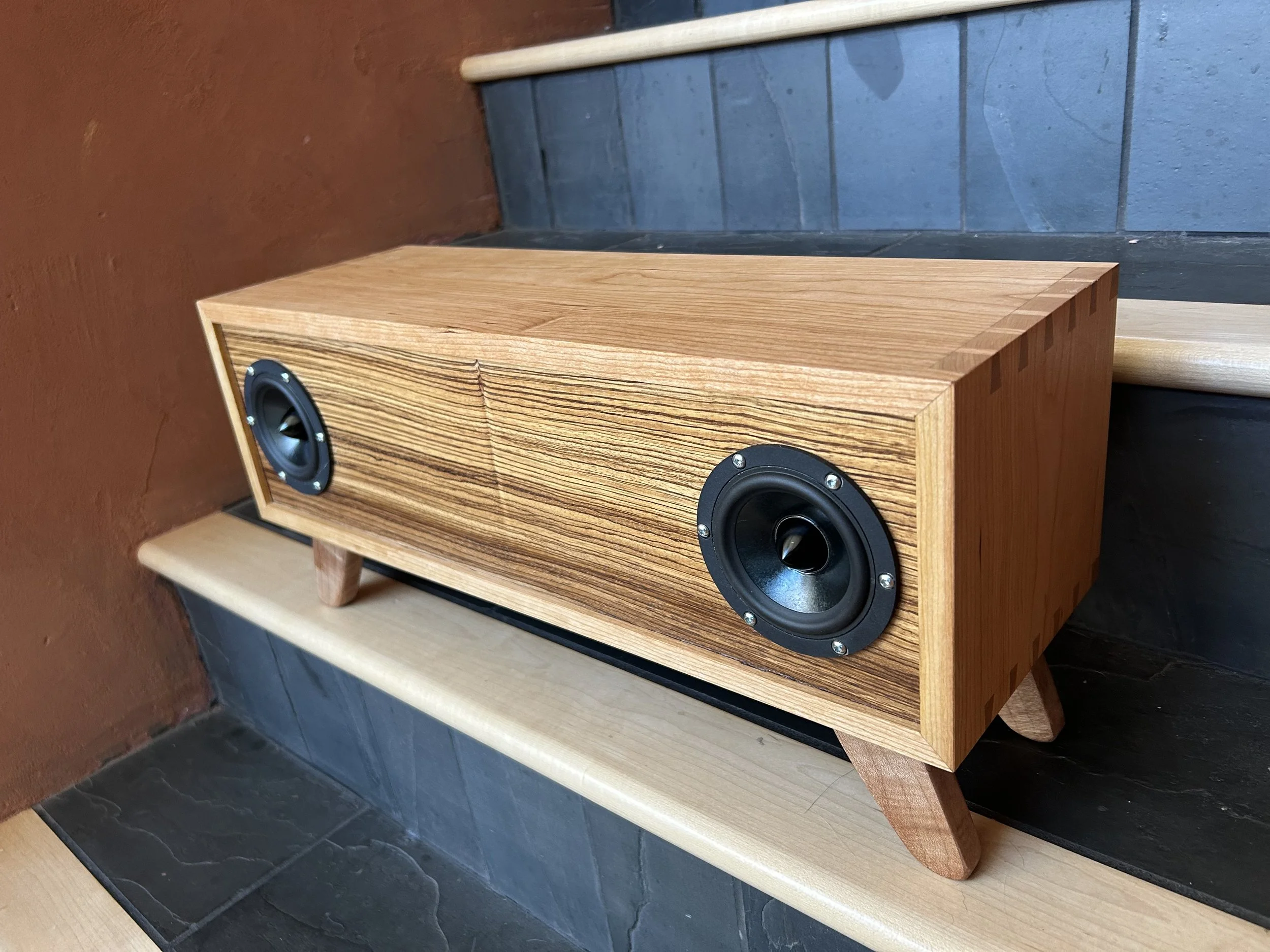 A wooden speaker with two black drivers is placed on a stepped wooden surface against a dark grey tiled wall and a brown wall.