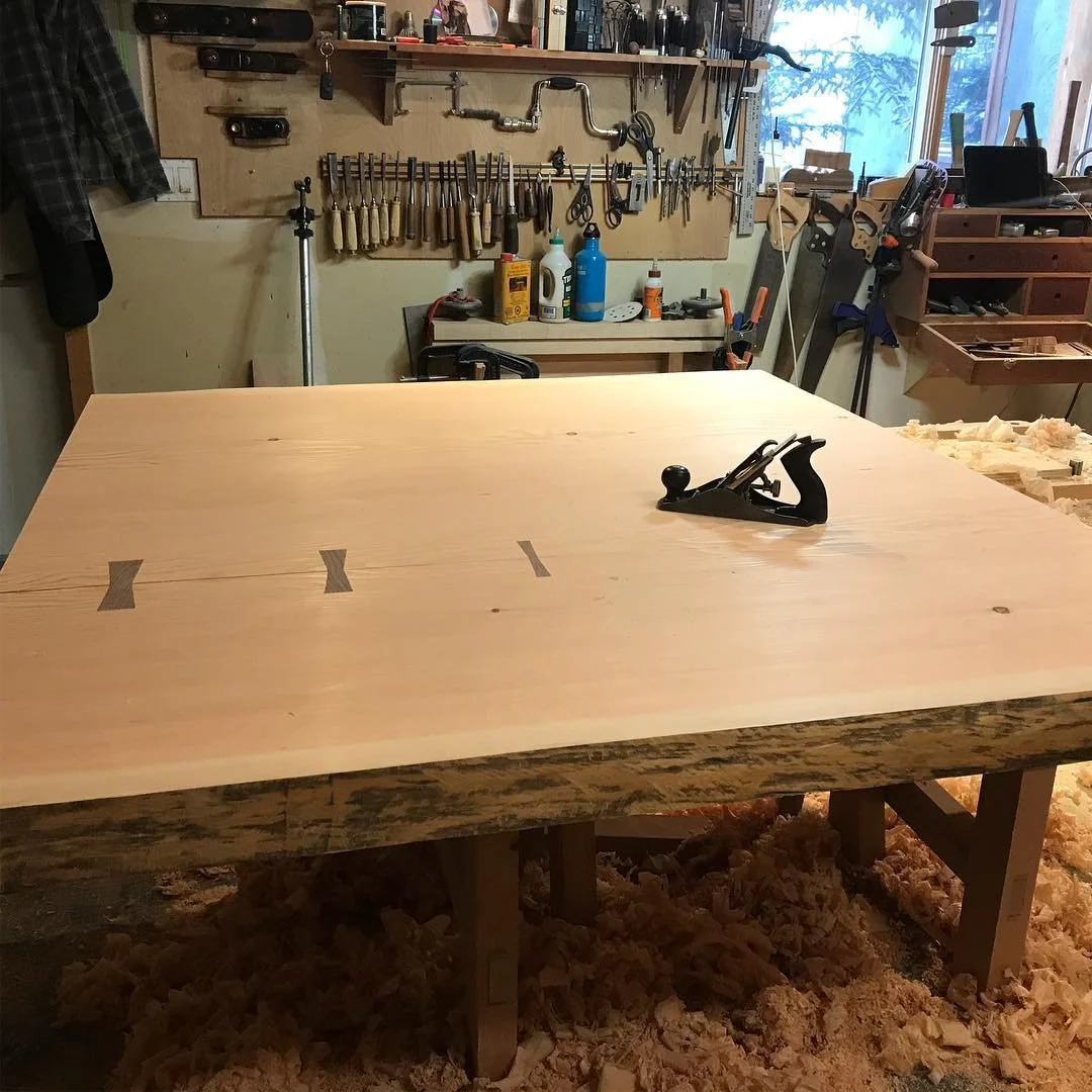 Woodworking shop with a large finished table in the center, clamps on the surface, and woodworking tools and supplies on the walls and shelves in the background.