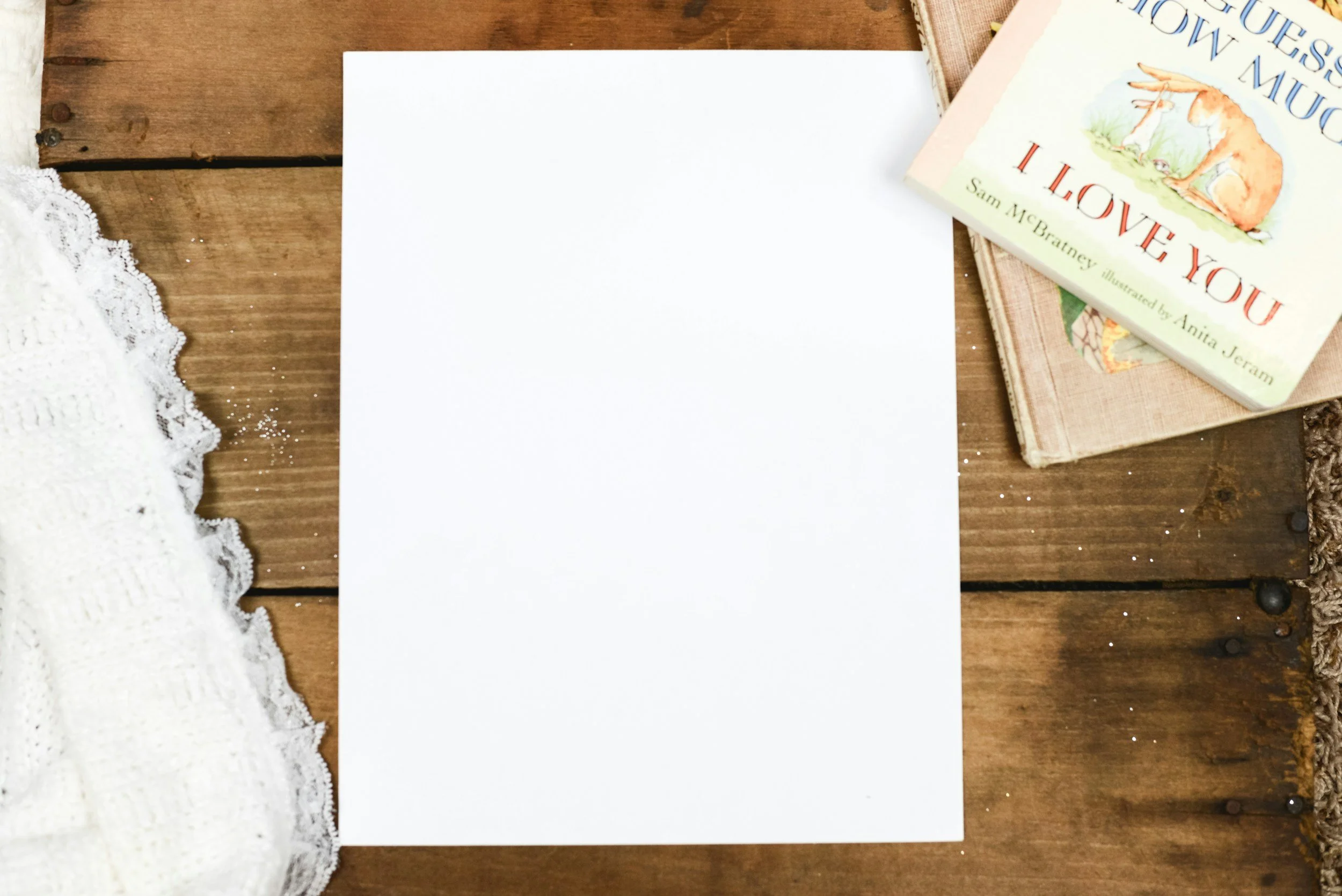 A flat lay image of a wooden table with a white piece of paper, a white knitted fabric, and three books stacked on top of each other, with the top book titled 'Mouse Houses of the Mouse' by Sam McBratney. The books have colorful illustrations.