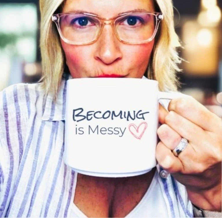A woman with blonde hair and glasses holding a white mug with the words 'BECOMING is Messy' and a pink heart, near her lips.