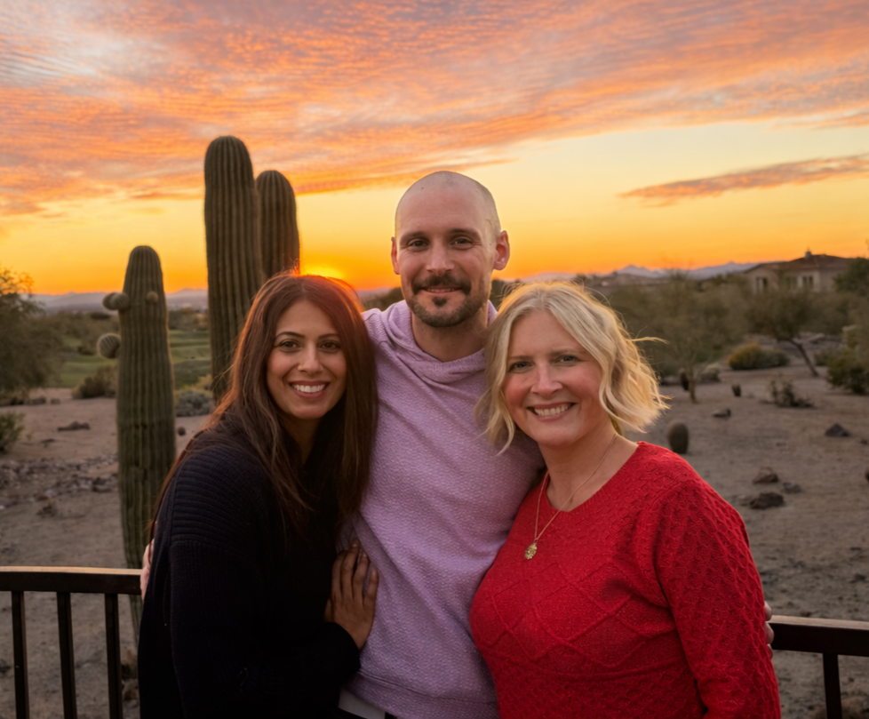 A group of three people smiling together outdoors at sunset, with desert landscape and cactus in the background.