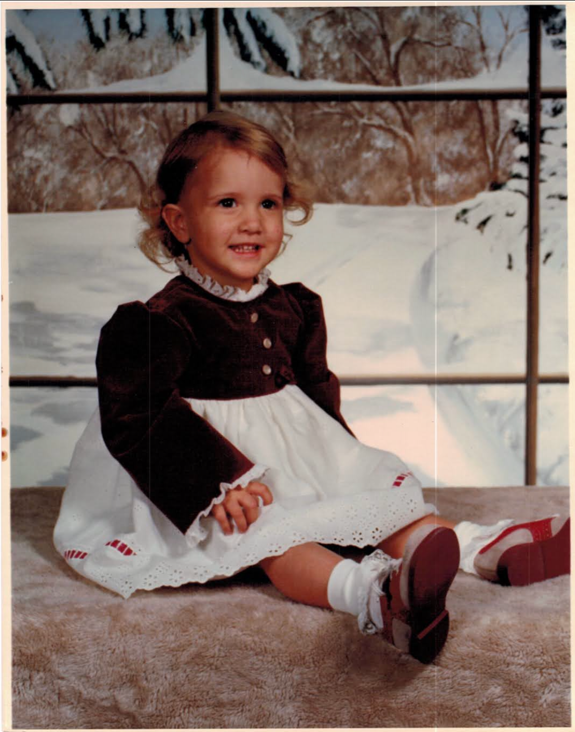 A young girl with curly hair sitting on a furry surface, smiling at the camera. She is wearing a dark brown dress with buttons and a white frilly apron, along with white socks and red shoes. The background shows a winter scene with snow-covered trees and a fence.