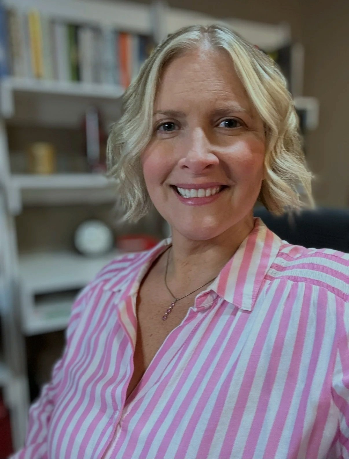 Portrait of a woman with blonde hair smiling, wearing a pink and white striped shirt, in a room with a bookshelf in the background.