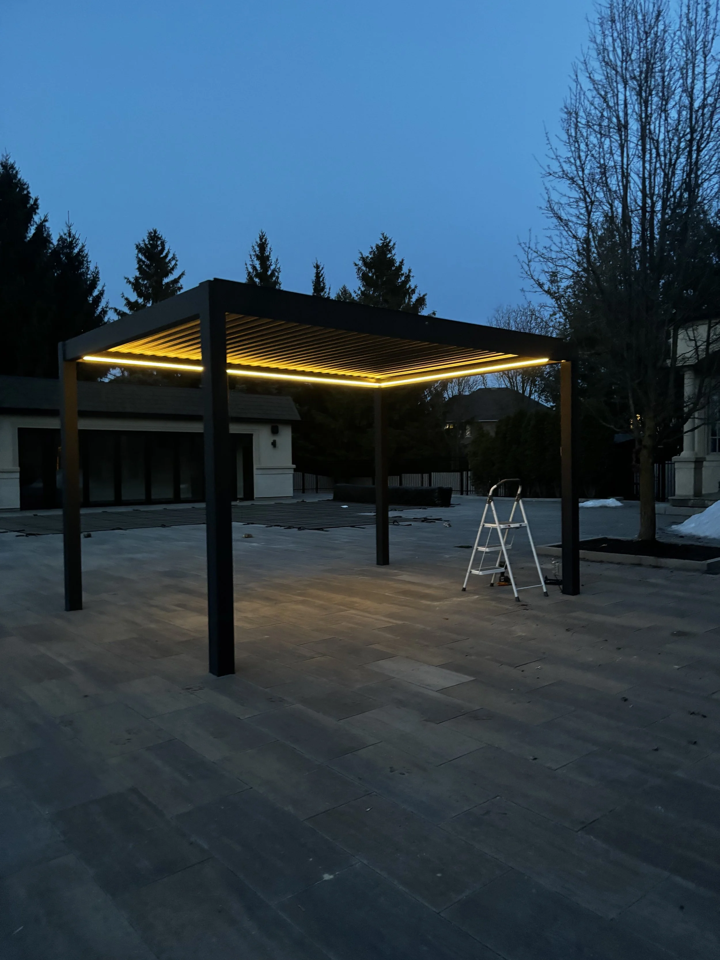 Empty outdoor patio area with a modern pergola illuminated by yellow lighting, trees in the background, and a step ladder on the tiled ground during dusk or early evening.