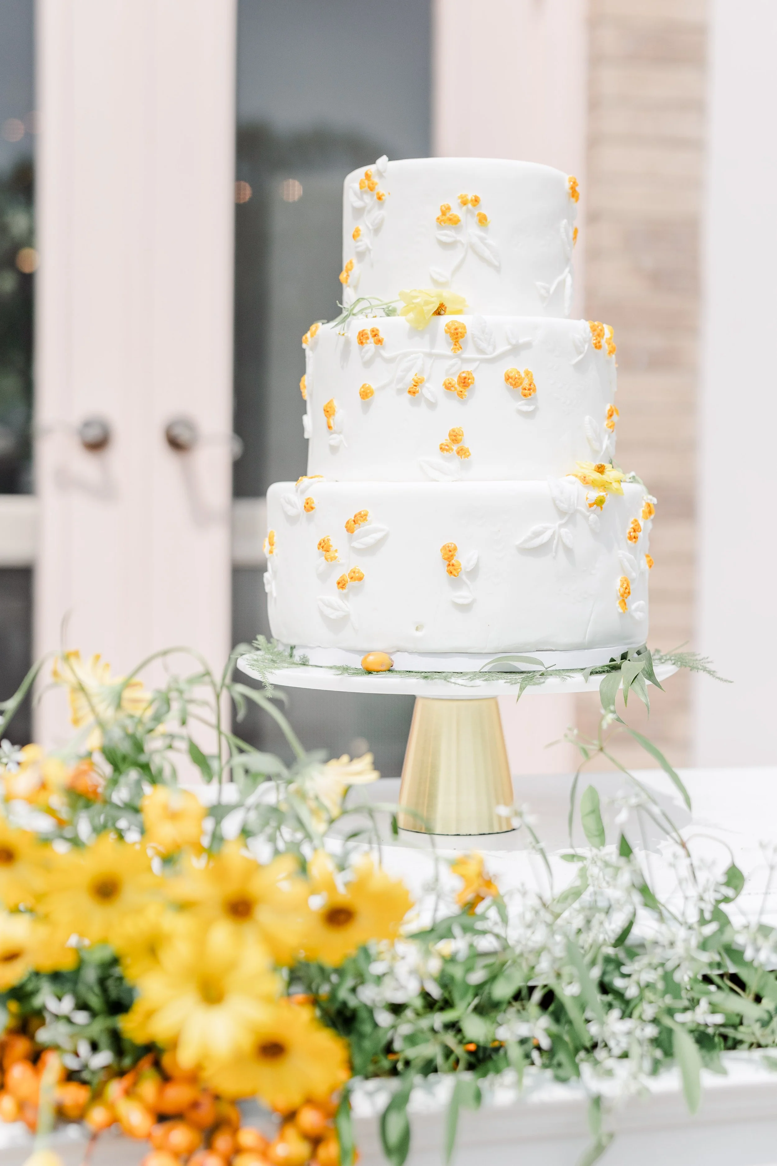 A three-tier white wedding cake decorated with small yellow flowers and white leaves, placed on a gold cake stand. Floral arrangements with yellow flowers and greenery are in the foreground.