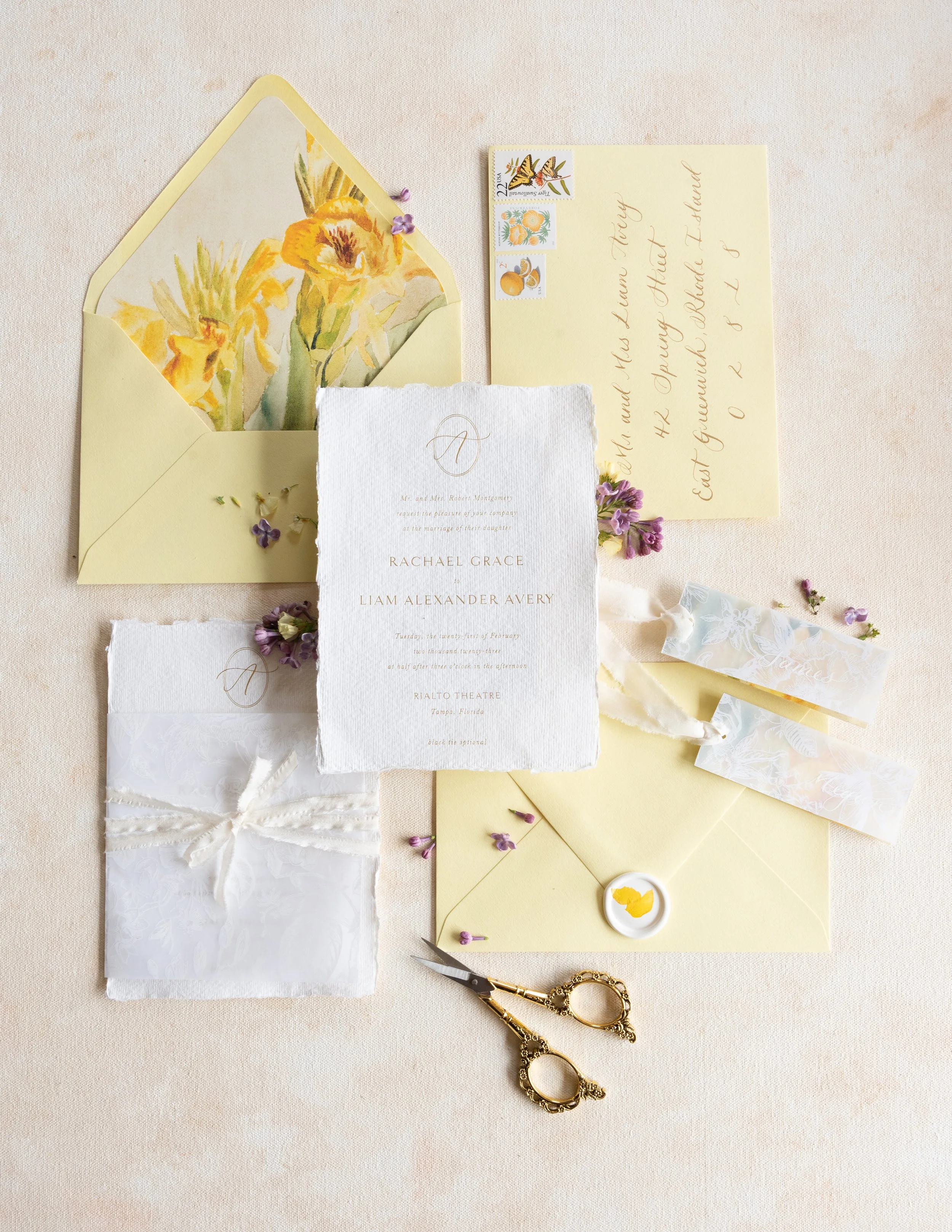 Wedding invitation suite with yellow envelopes, floral watercolor inside envelope, handwritten address on yellow card, torn textured RSVP card, lace favor pouch with ribbon, small purple flowers, and gold scissors on a neutral fabric surface.