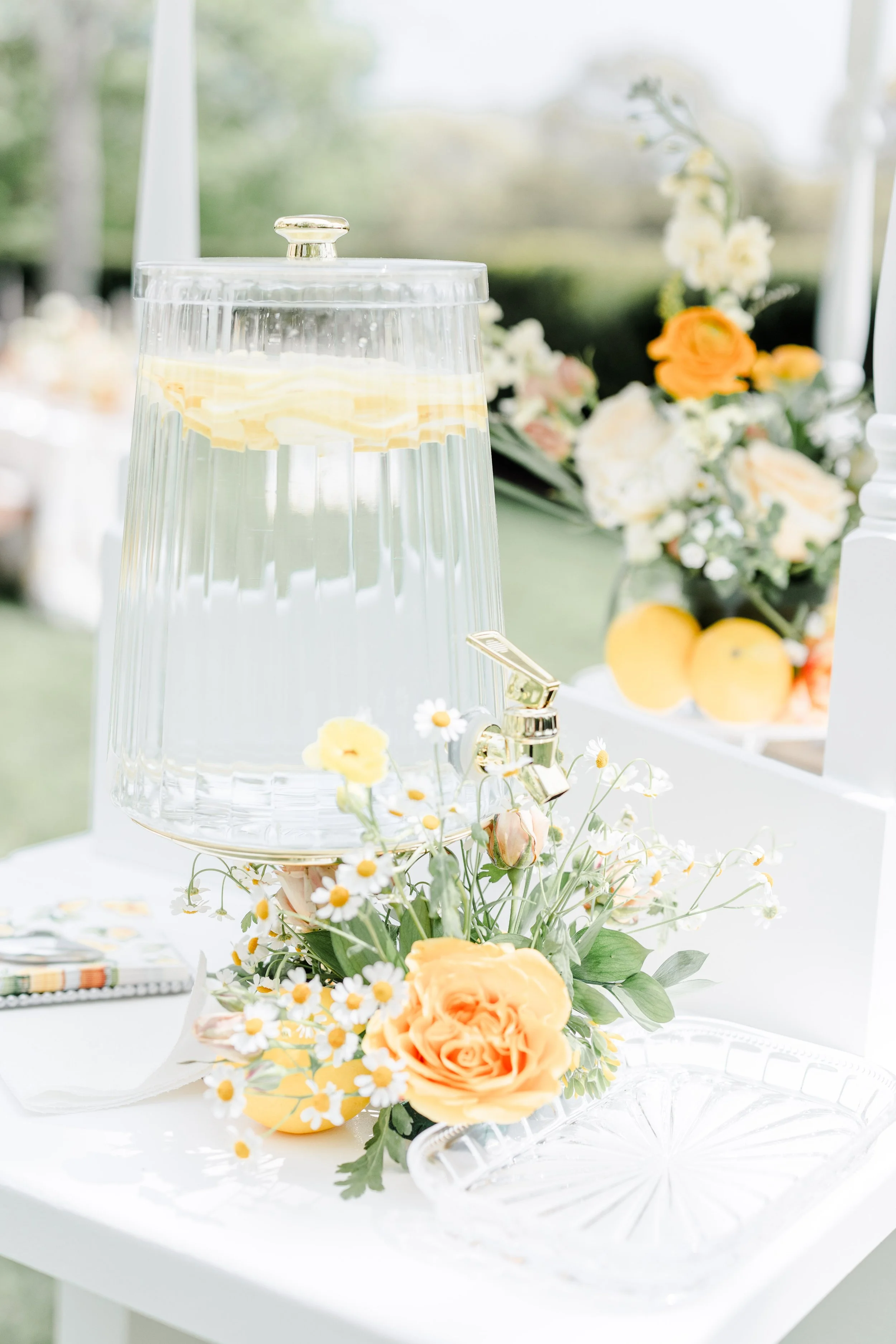 A glass drink dispenser filled with water and lemon slices, surrounded by a floral arrangement of yellow roses, white daisies, and greenery on a white table with a blurred outdoor background.